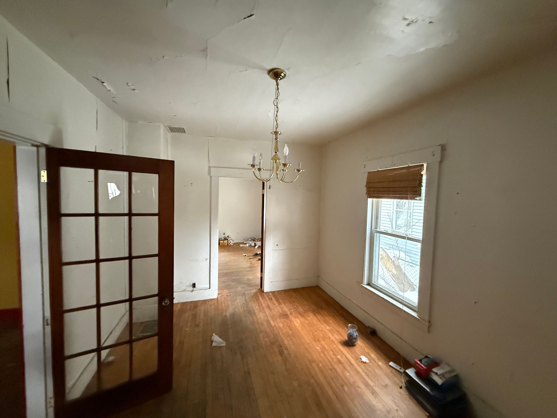 An empty room with light wood floors, a hanging chandelier, a window with a brown shade, and an open glass-paneled door.