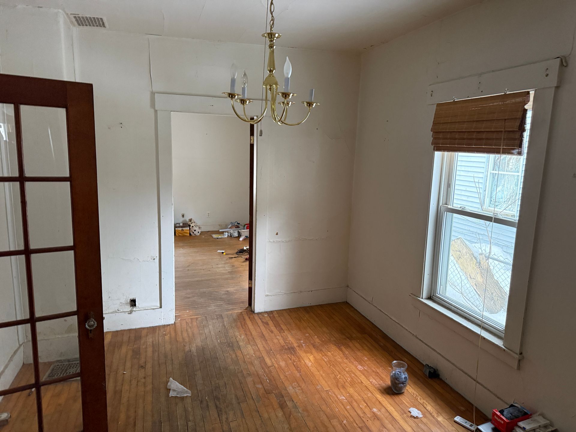A vacant room with hardwood floors, white walls, a brass chandelier, a wood-framed door, and a window with a bamboo shade.