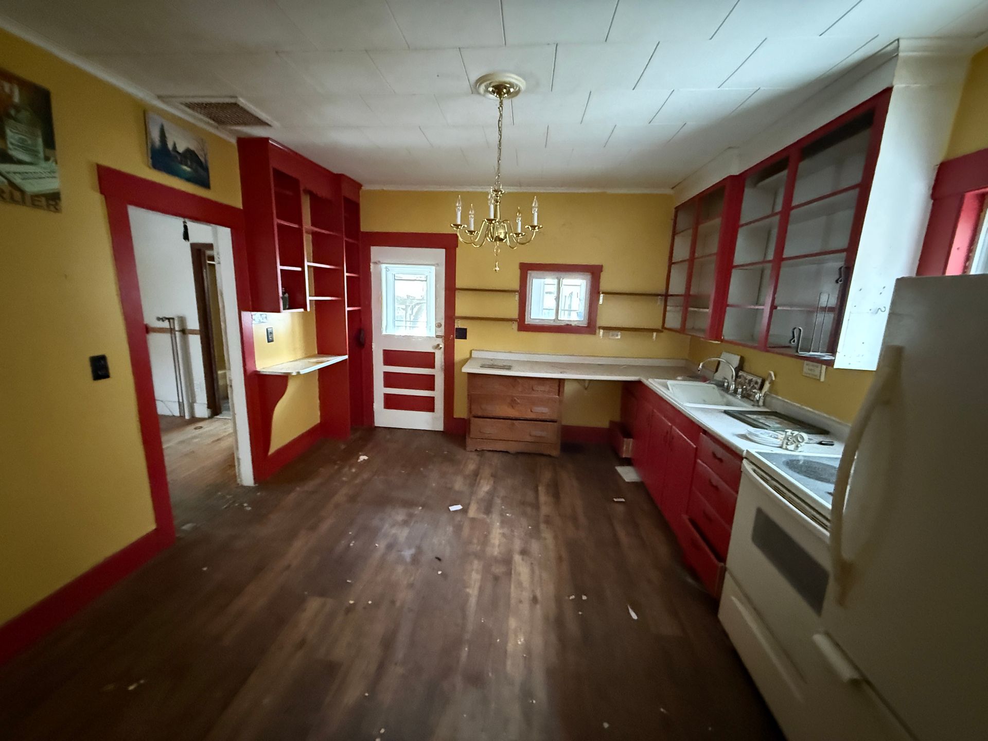 A bright kitchen with yellow walls, red trim, dark wood-look floors, a white refrigerator, and a gold chandelier.