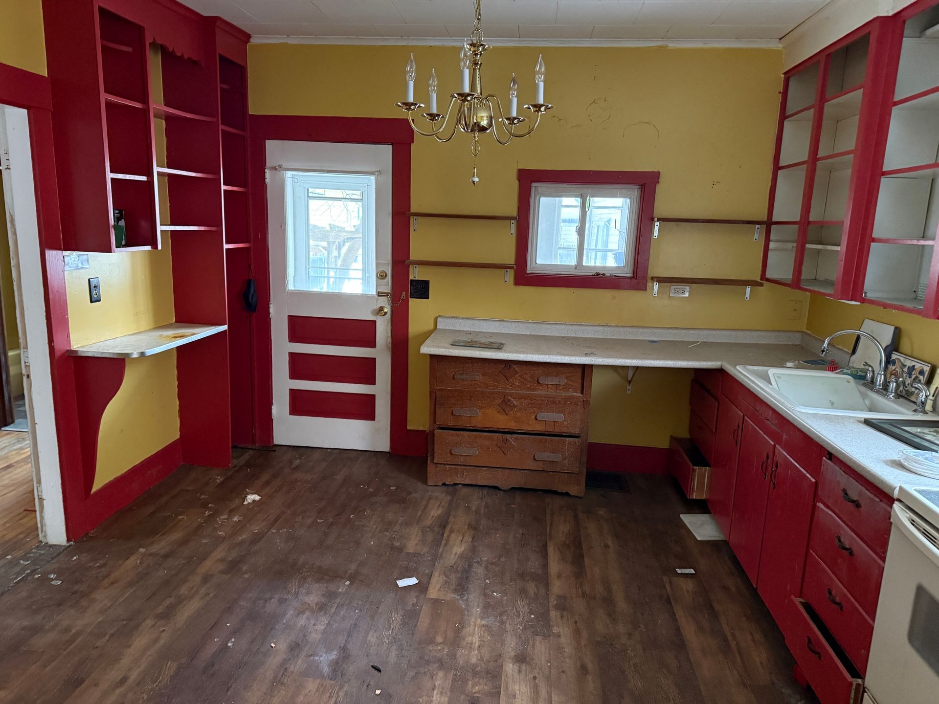 A kitchen with yellow walls, red cabinets, wood flooring, a central door, and a white sink countertop.