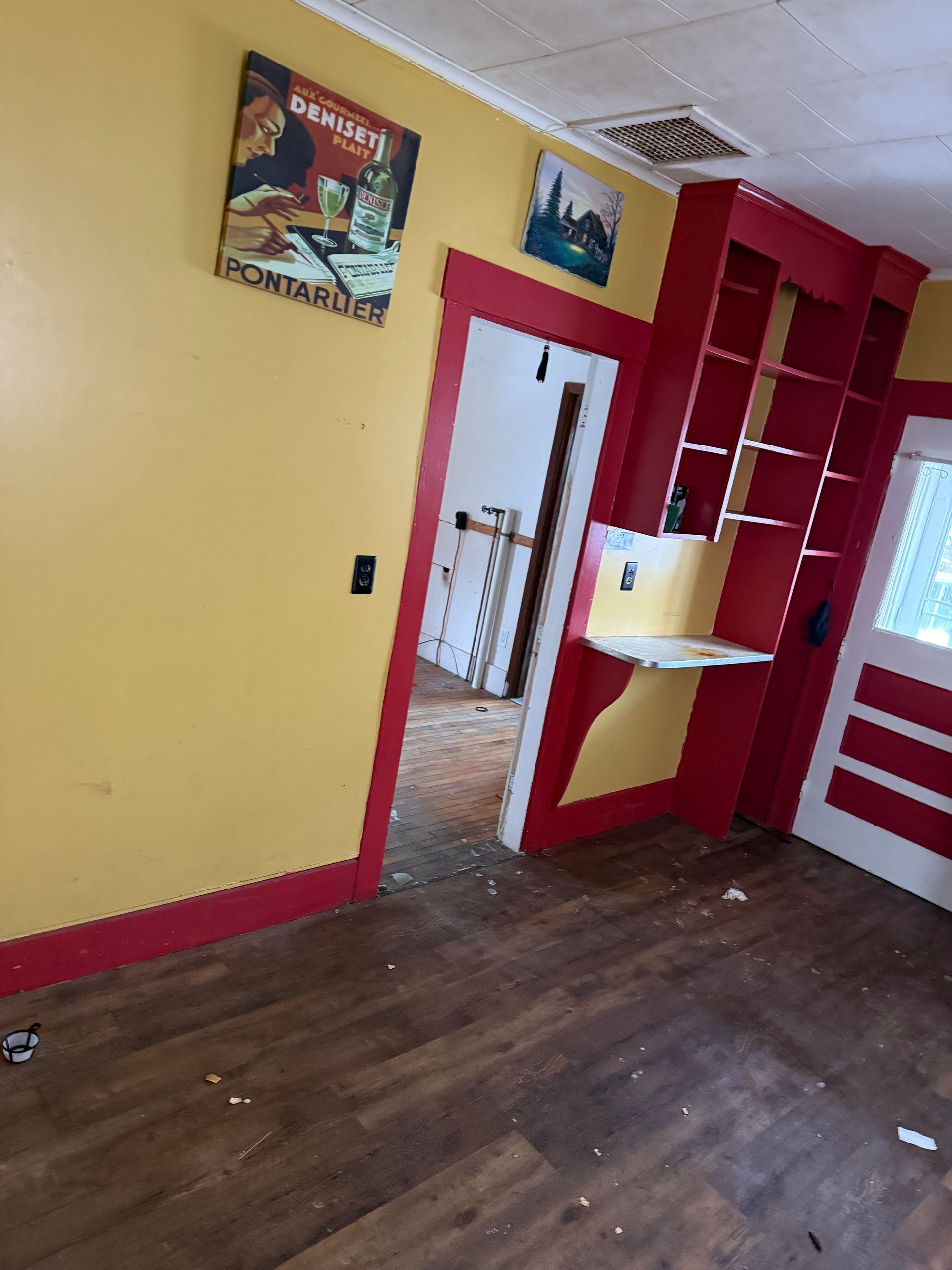 Yellow room with red trim, hardwood floors, a wall shelf, a doorway to another room, and two pieces of wall art.