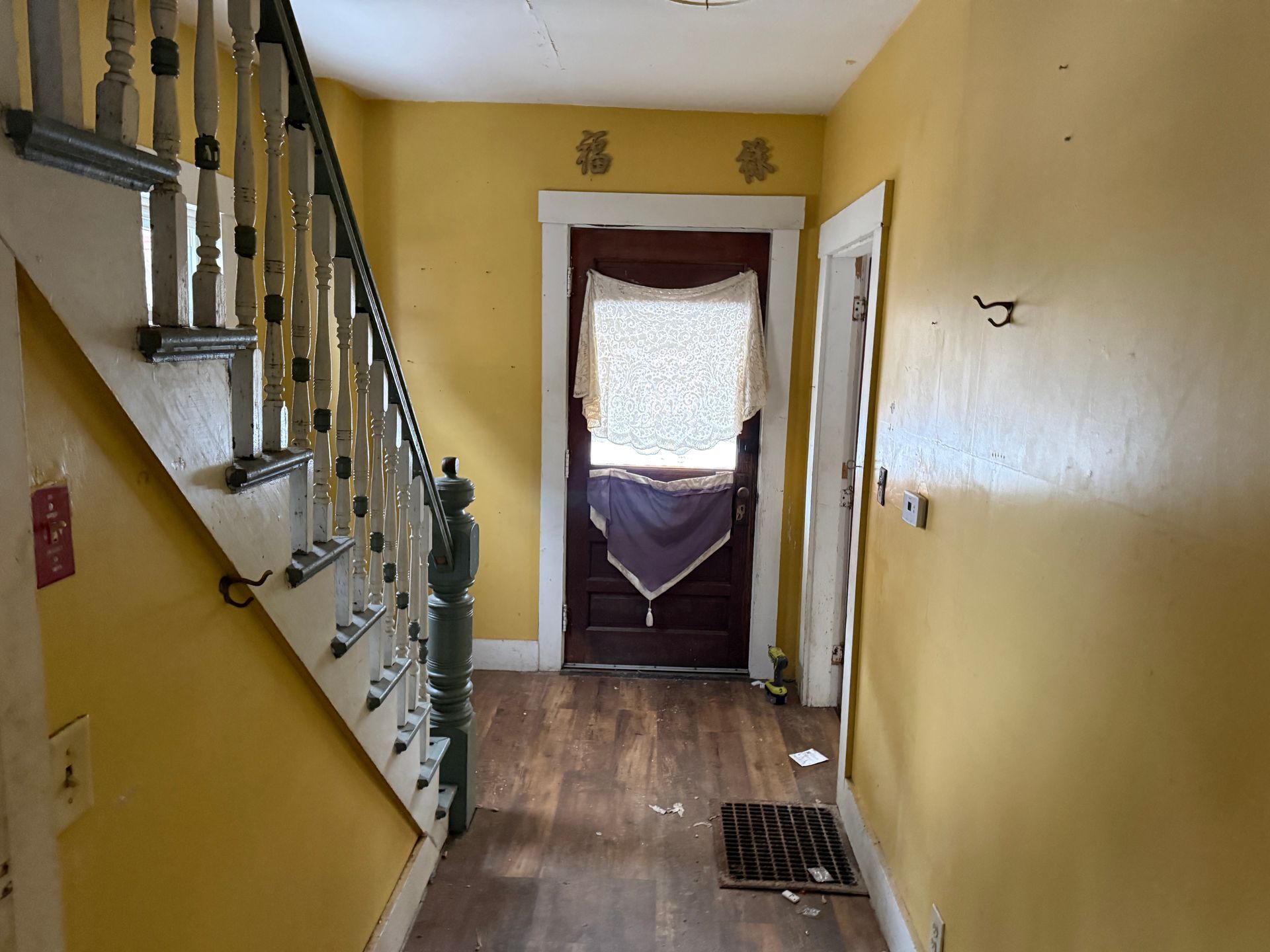 A yellow entryway with a wooden staircase on the left, a dark front door with a lace curtain, and a wood-look floor.