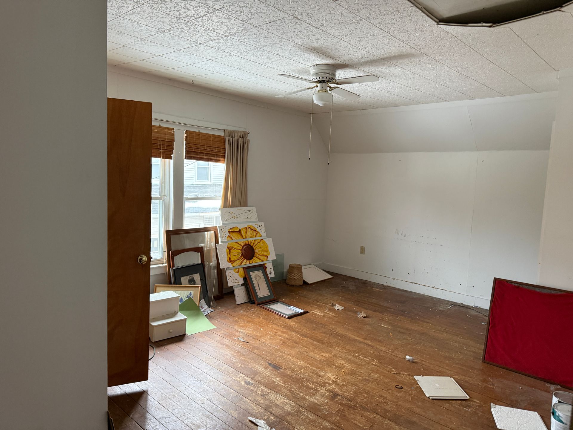 An empty room with hardwood floors, a ceiling fan, a window with curtains.