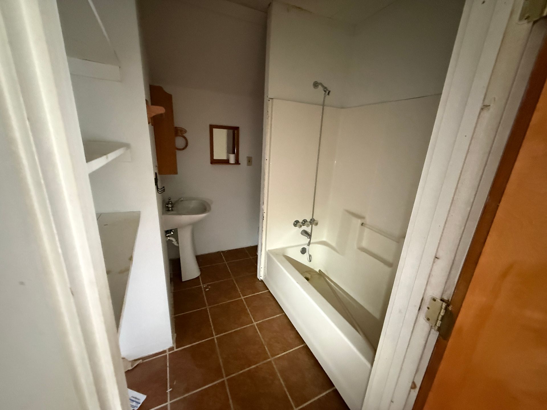 A bathroom with white walls, brown tiled floors, a white pedestal sink, and a bathtub with a shower fixture.