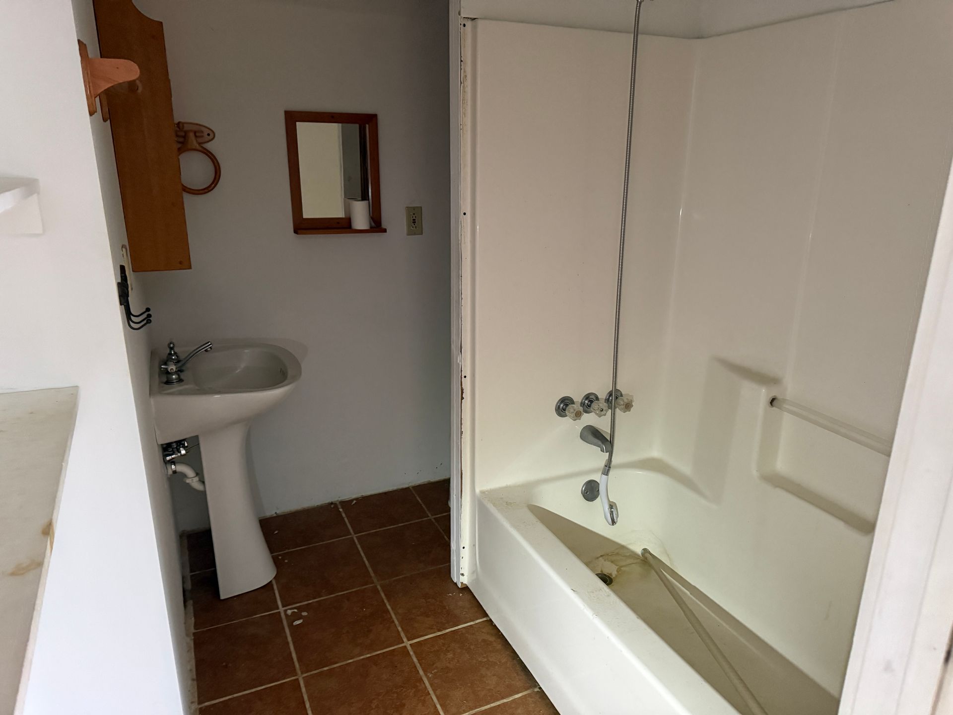 A bathroom with a pedestal sink, a small wall mirror, and a white bathtub-shower unit on a brown tiled floor.