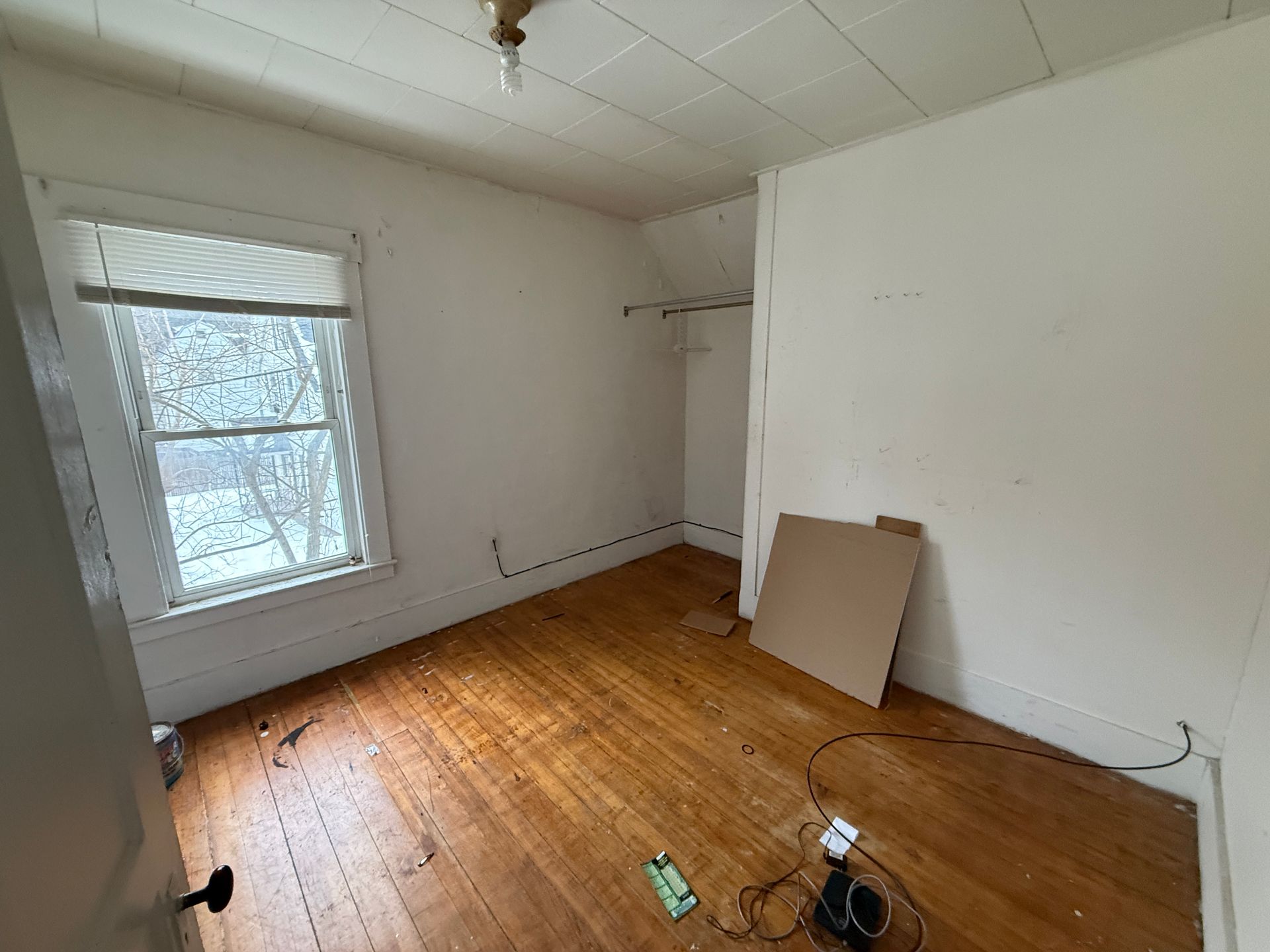 An empty room with white walls, a single window looking out on snow, hardwood floors, and a small built-in closet area.