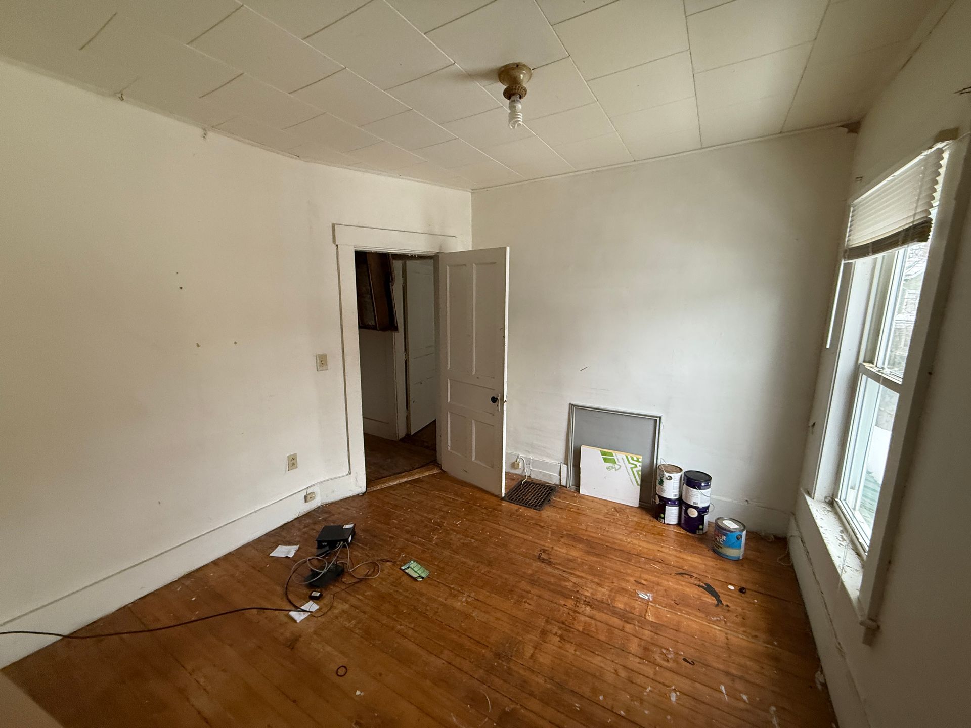 An empty room with white walls, wooden floors, a doorway, a ceiling light, and a window with a blind.