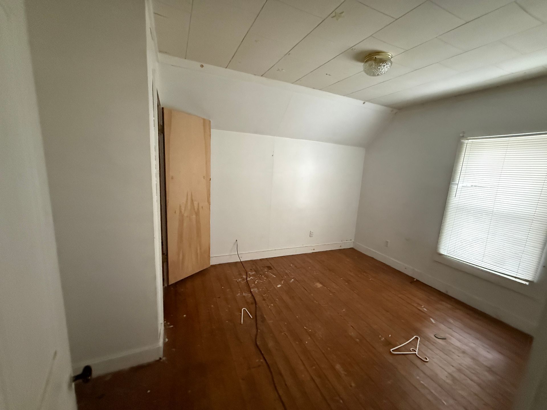 An empty room with white walls, a sloping ceiling, wooden floors, a light-colored wooden door, and a window with blinds.