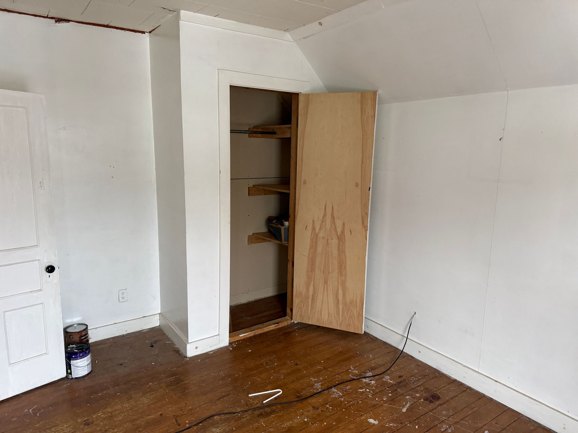 A room with white walls, a wooden floor, a closed white door, and an open closet with wooden shelves and a plywood door.