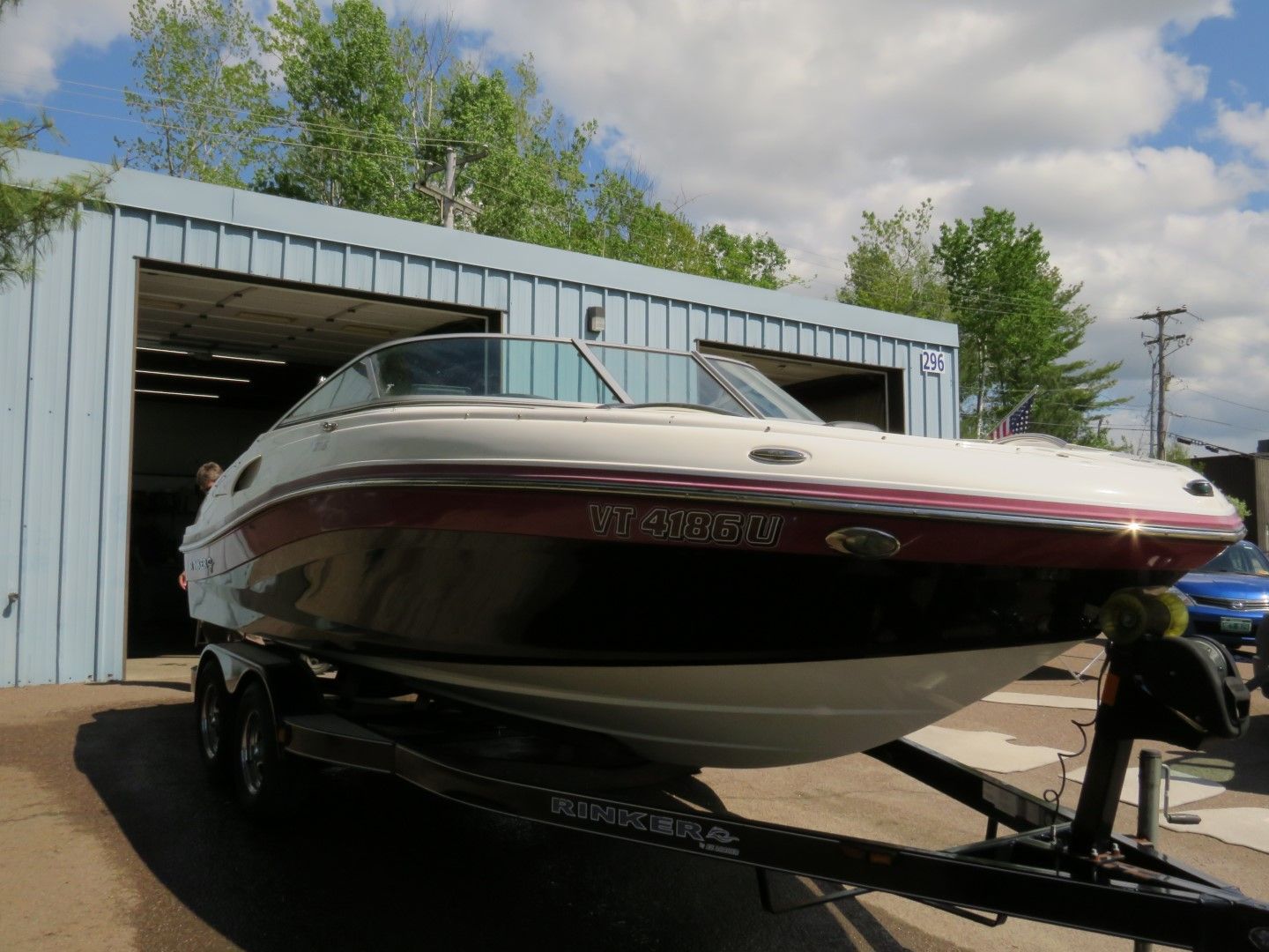 White and maroon boat on a trailer, in front of a blue garage, sunny day.
