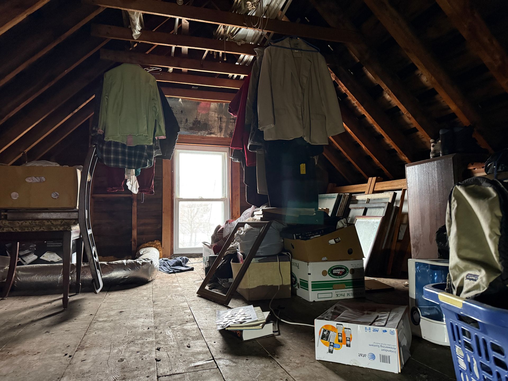An attic with exposed wooden rafters, hanging clothes, cardboard boxes, and a central window letting in light.