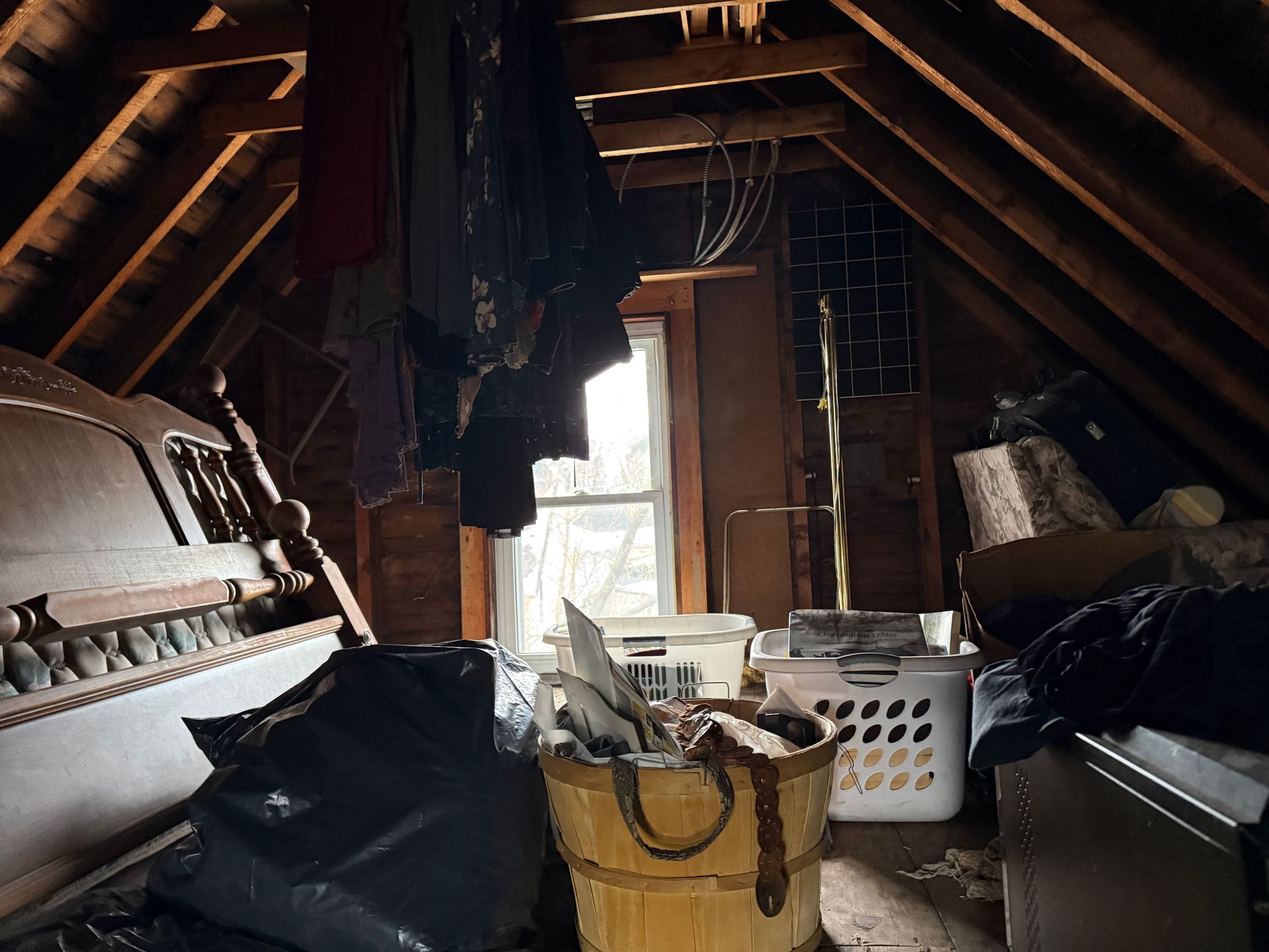 An attic interior features a wooden bed frame, hanging clothing, a storage basket, and laundry bins near a small window.