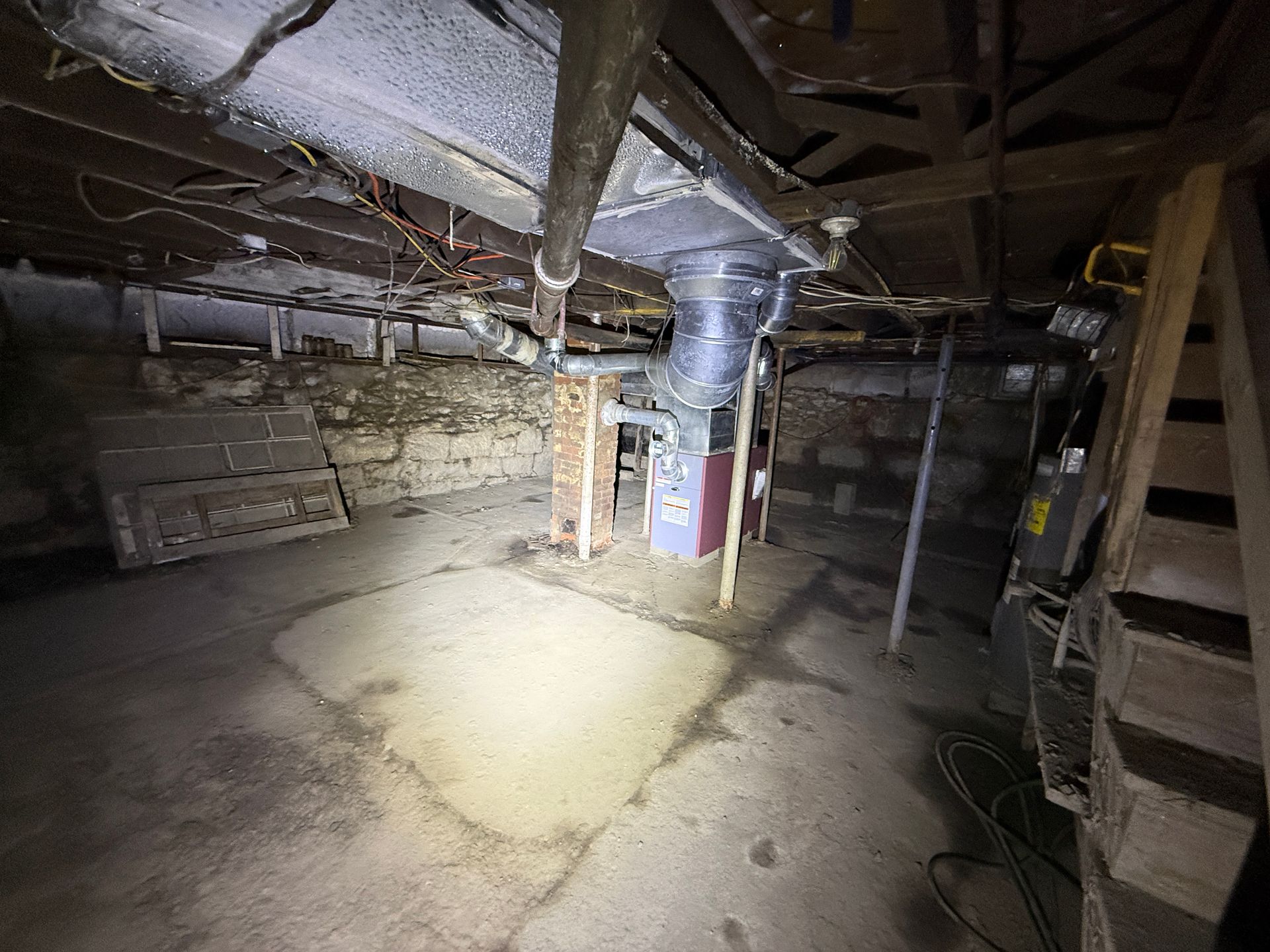 A basement with a stone foundation, wooden floor joists, a furnace, and stairs on the right.