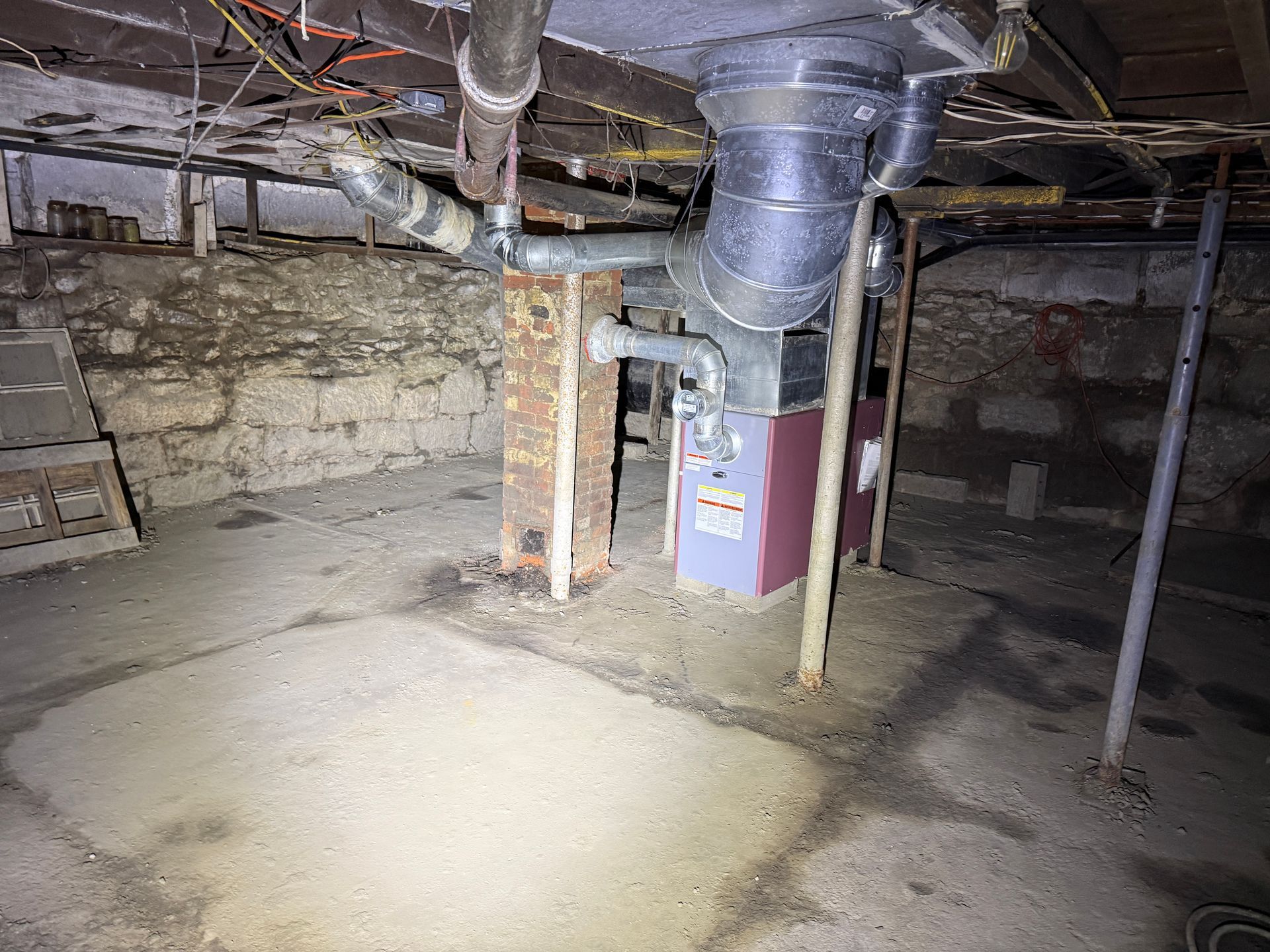 A furnace sits in a basement with stone walls, concrete floors, and exposed ductwork supported by a metal post.