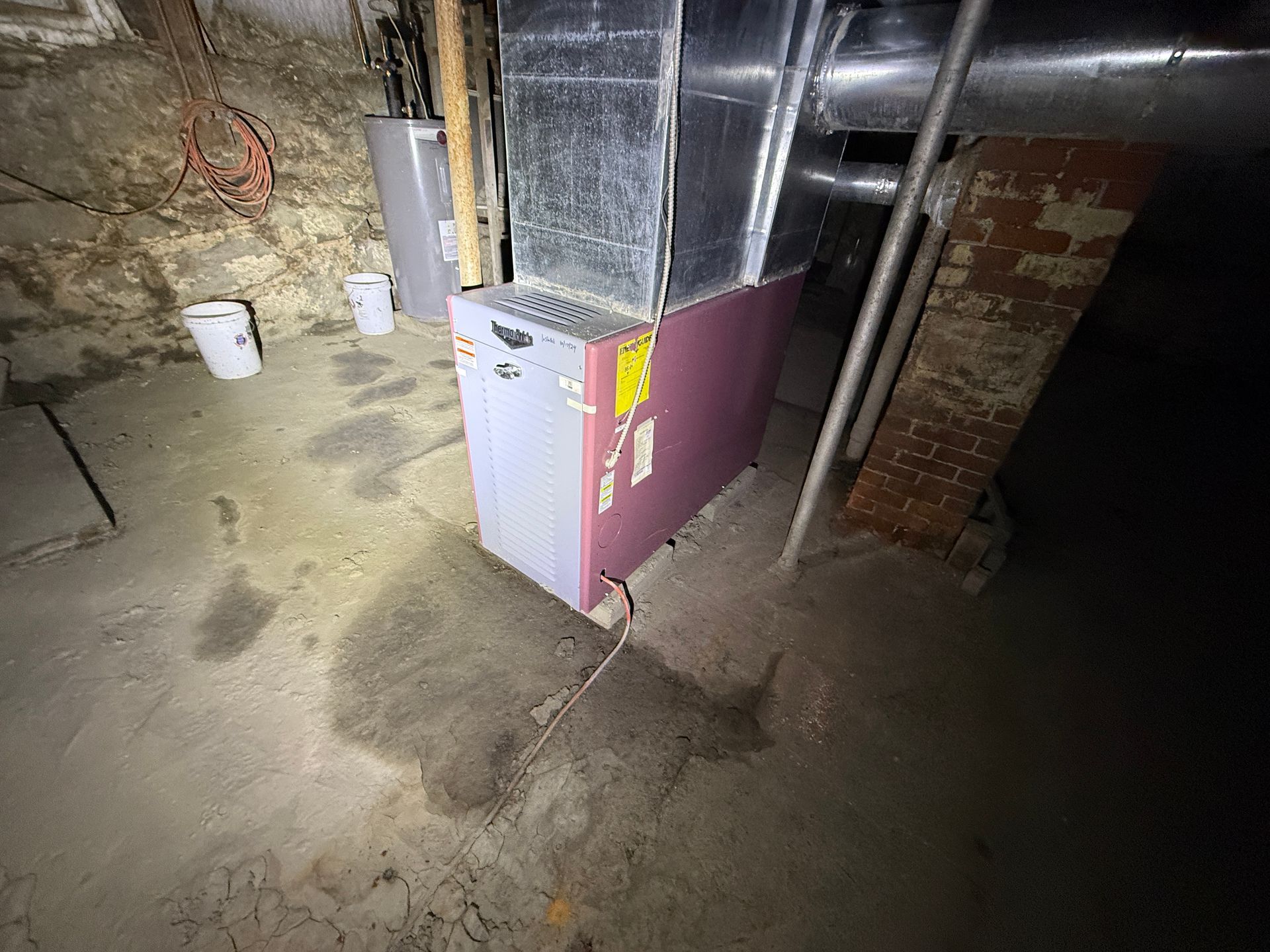 A red furnace and metal ductwork stand on a concrete basement floor near a brick chimney and water heater.