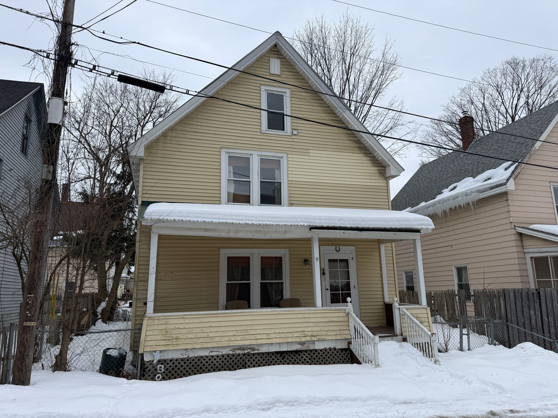 A two-story, yellow house with a covered front porch, seen in winter with snow on the ground and roof.