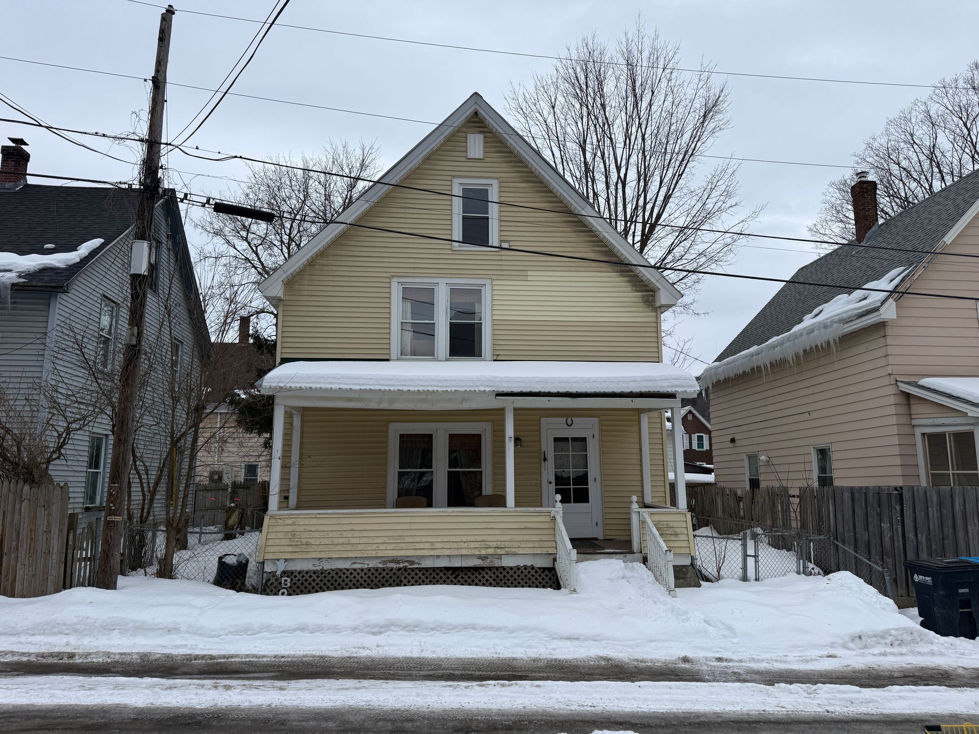 A two-story, yellow house with a front porch, covered in snow on a cloudy day, positioned between two other homes.