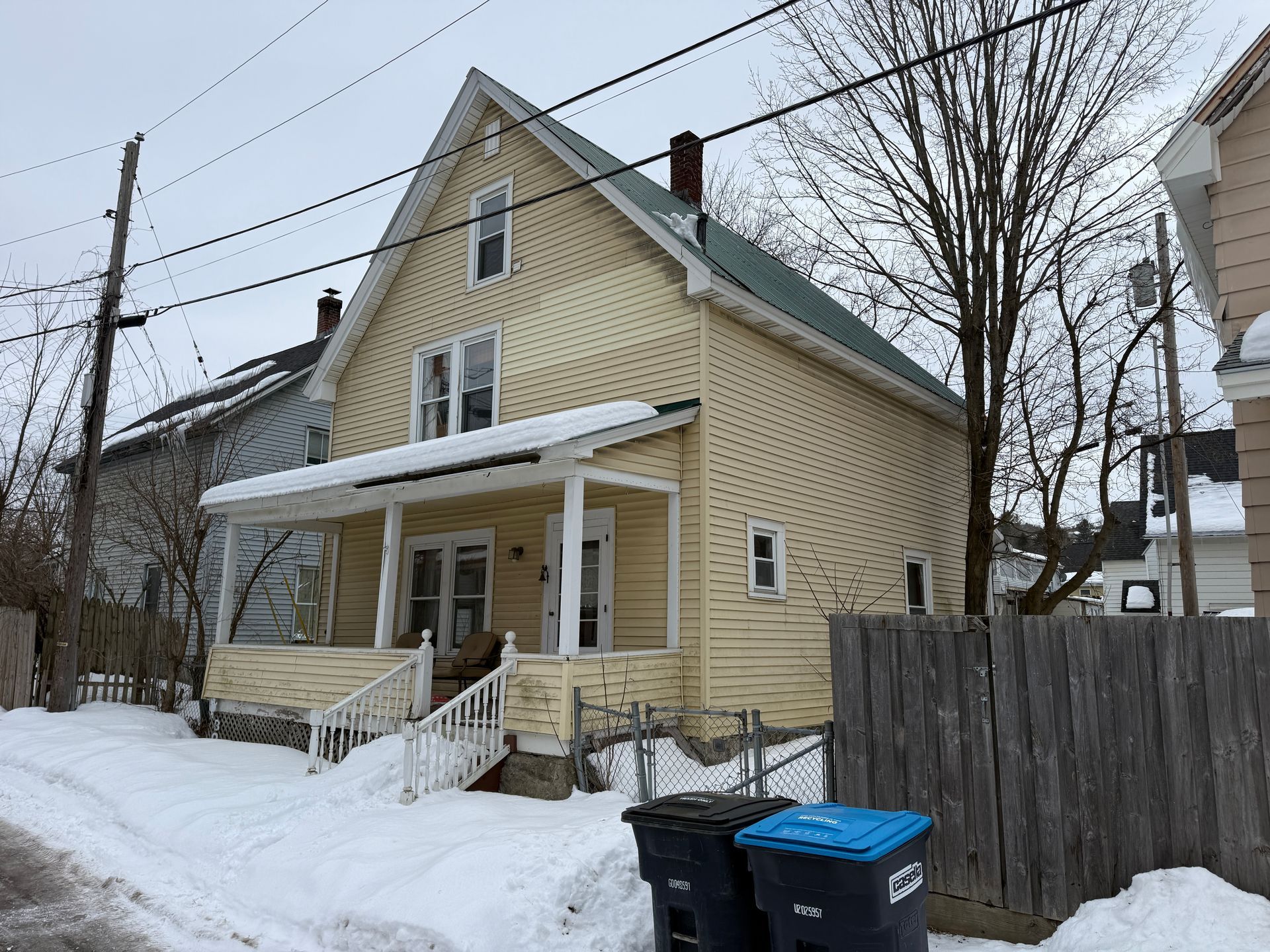 A pale yellow two-story house with a covered front porch, surrounded by snow in a residential neighborhood.
