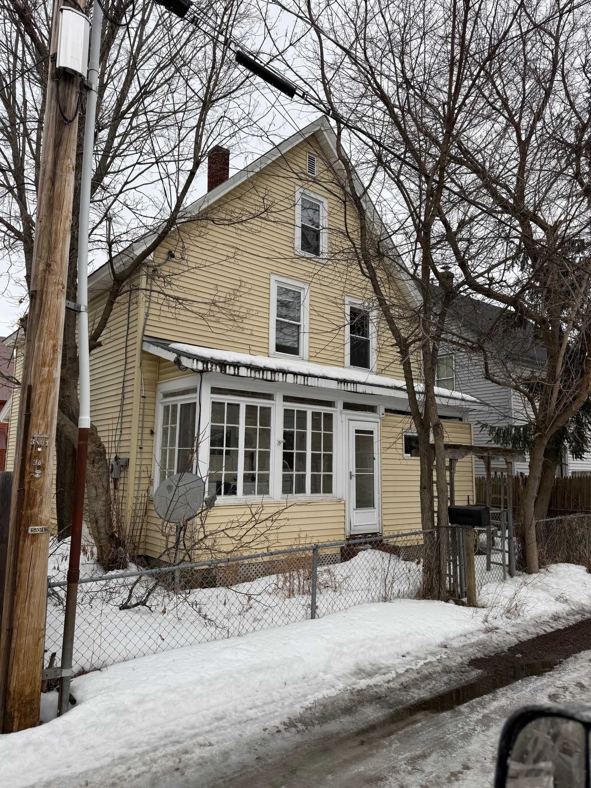 A two-story, pale yellow house with a white-framed enclosed porch, set behind a chain-link fence during winter.