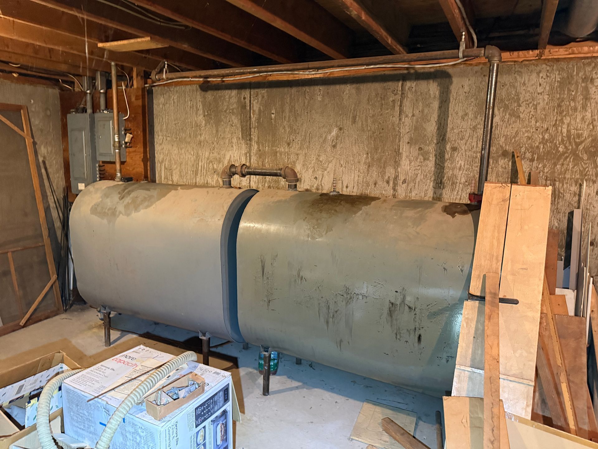 A horizontal oil tank in a basement with pipes, wires, and wooden beams.
