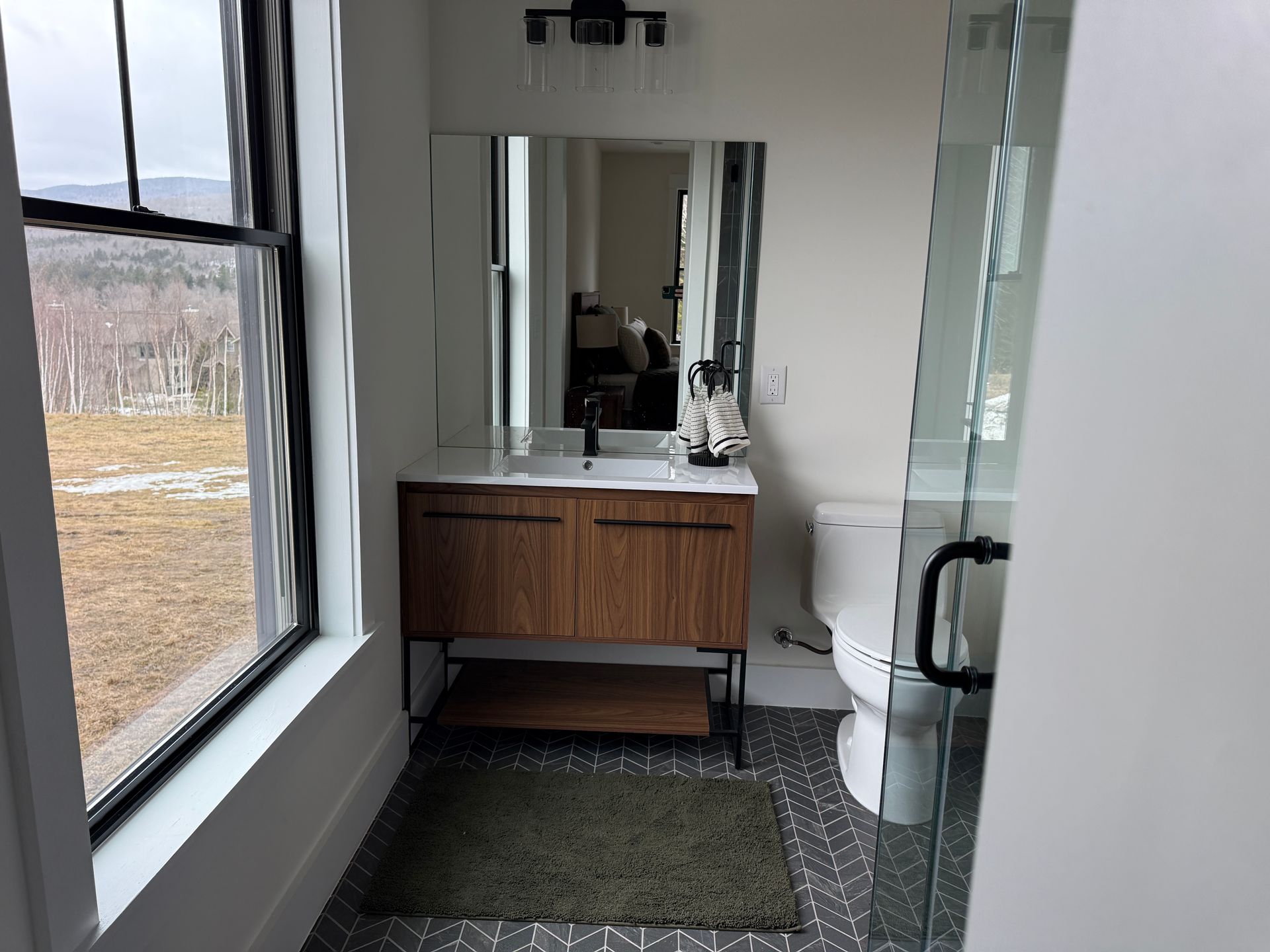 A modern bathroom with a wooden vanity, square sink, mirror, wall lighting, toilet, and a glass-enclosed shower.