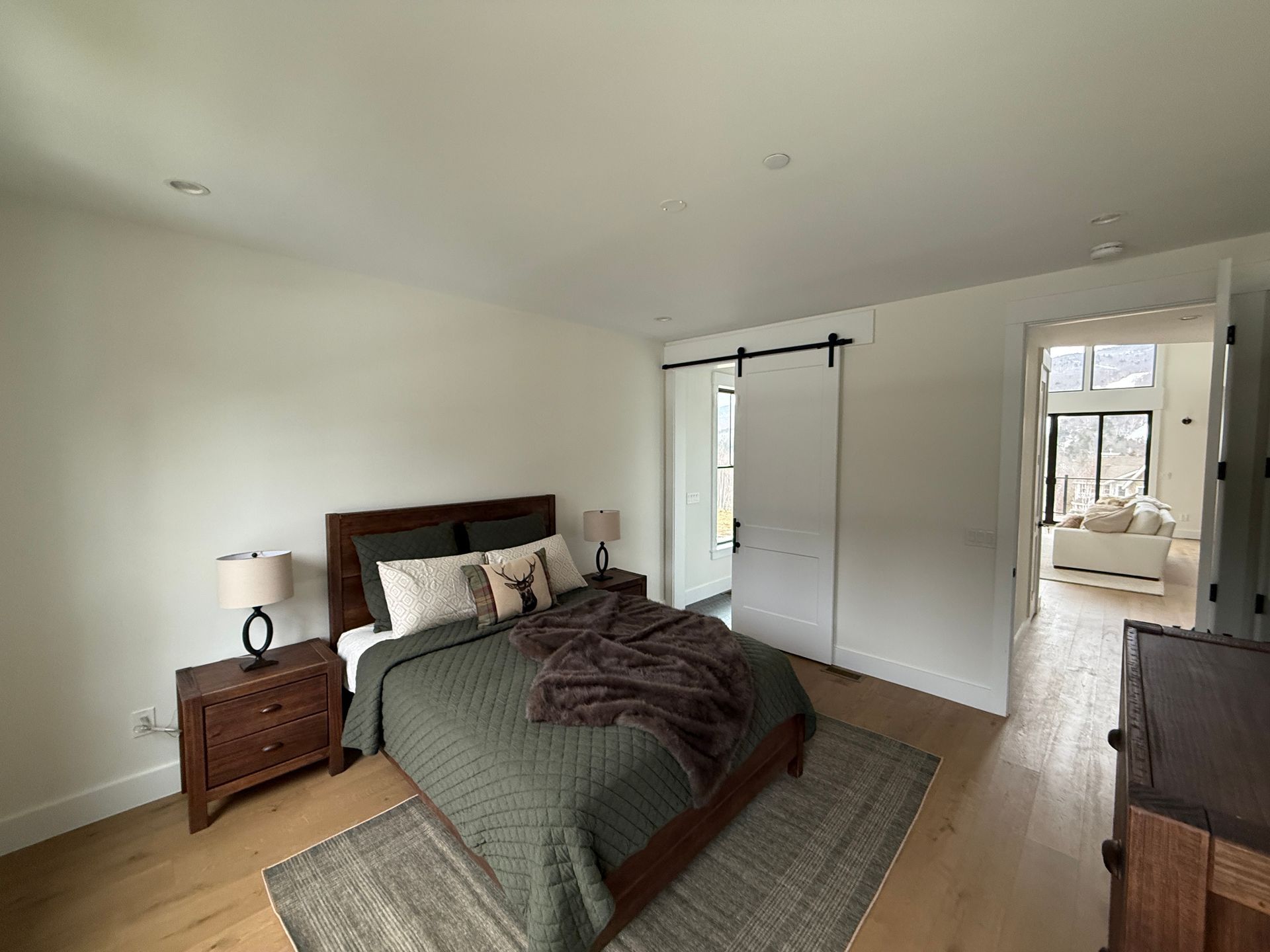 A cozy bedroom with a dark wooden bed, olive green quilt, matching nightstands, and a white barn door on light floors.