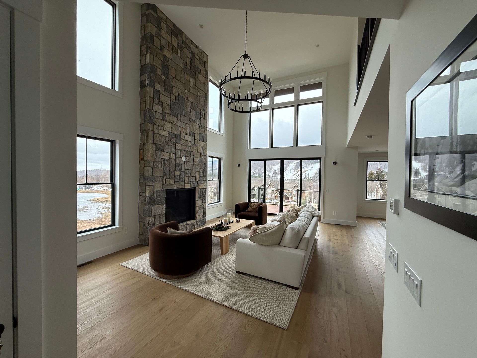 A spacious, modern living room with high ceilings, a stone fireplace, large windows, and neutral-toned furniture.