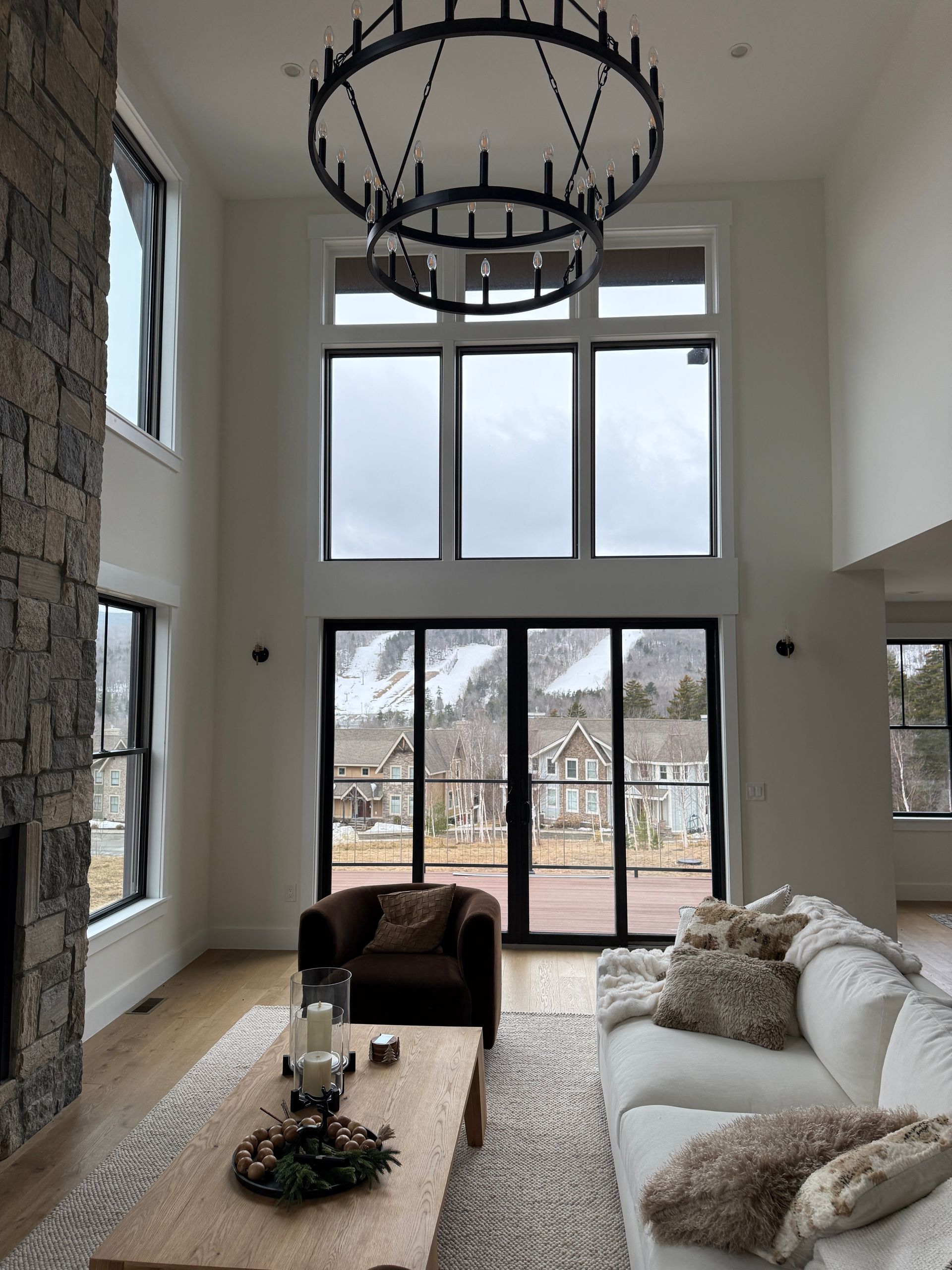 Living room with a stone fireplace, white sofa, dark armchair, and large windows revealing a snowy mountain landscape.