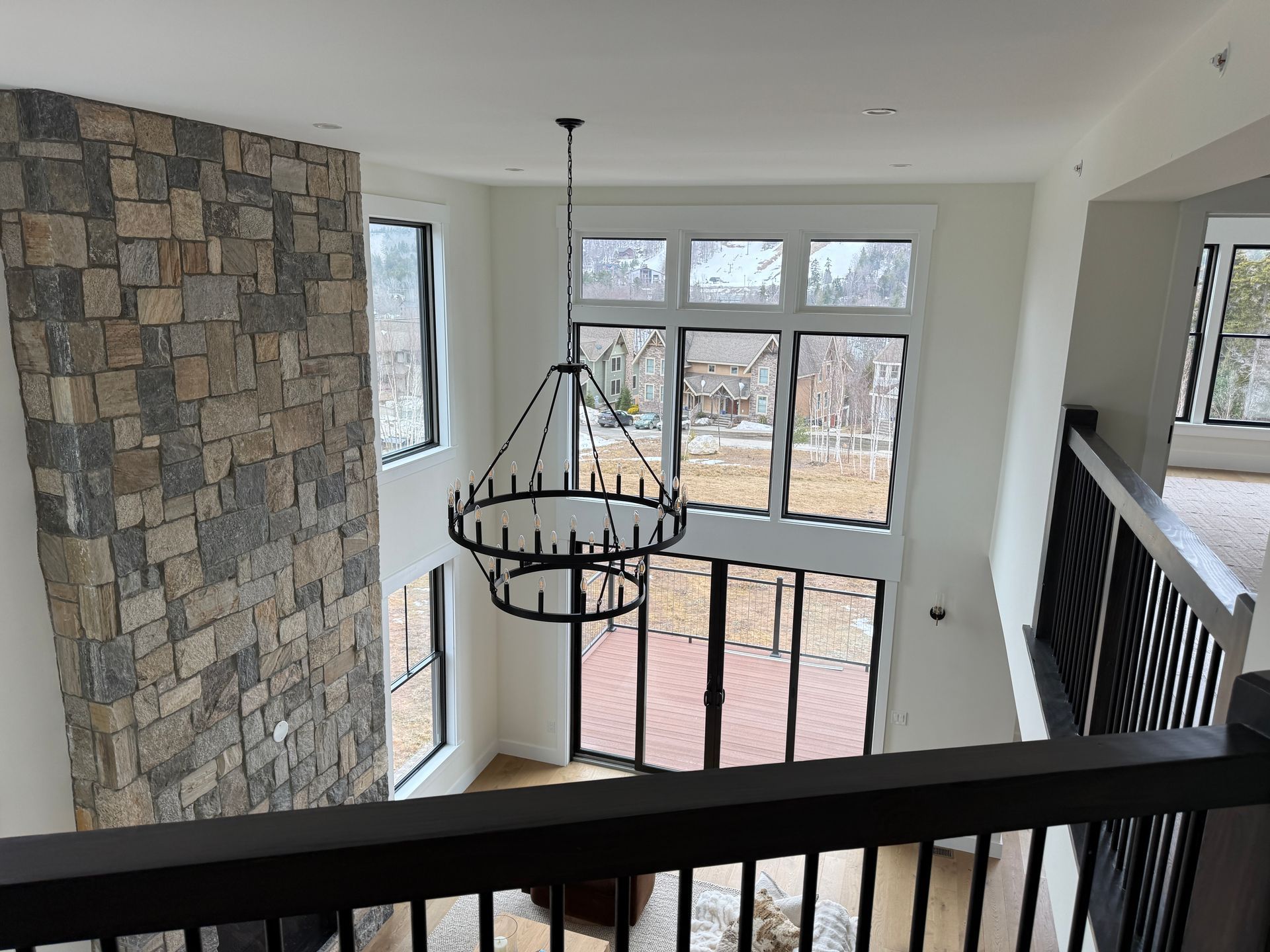 A tall stone fireplace overlooks a living area with floor-to-ceiling windows, a large round chandelier, and a railing.