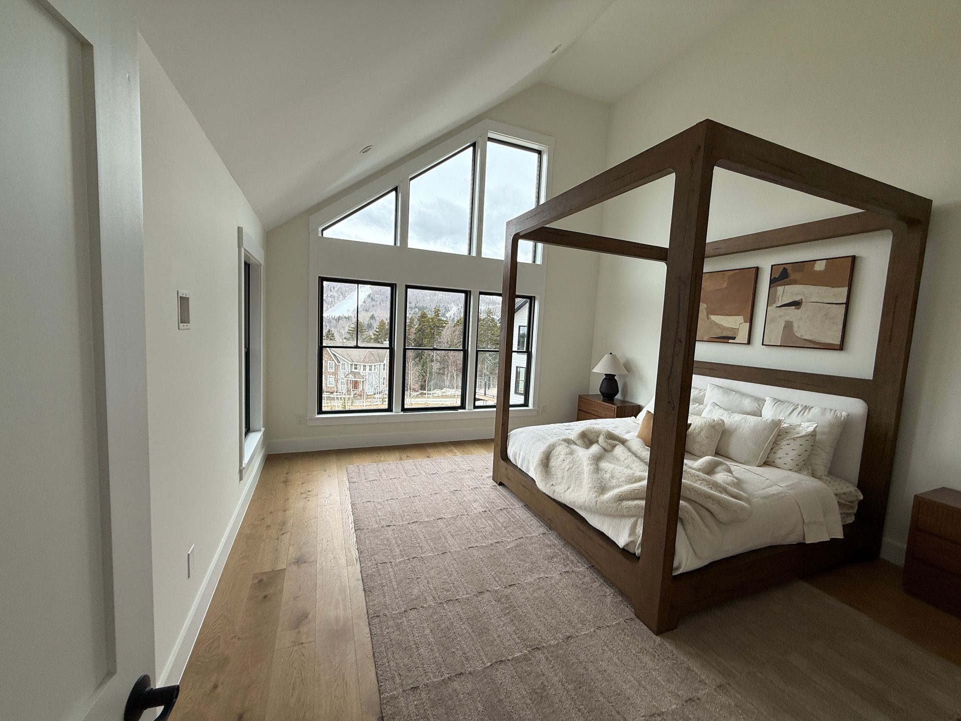 A modern bedroom with a wooden four-poster bed, large windows with mountain views, and light hardwood floors.