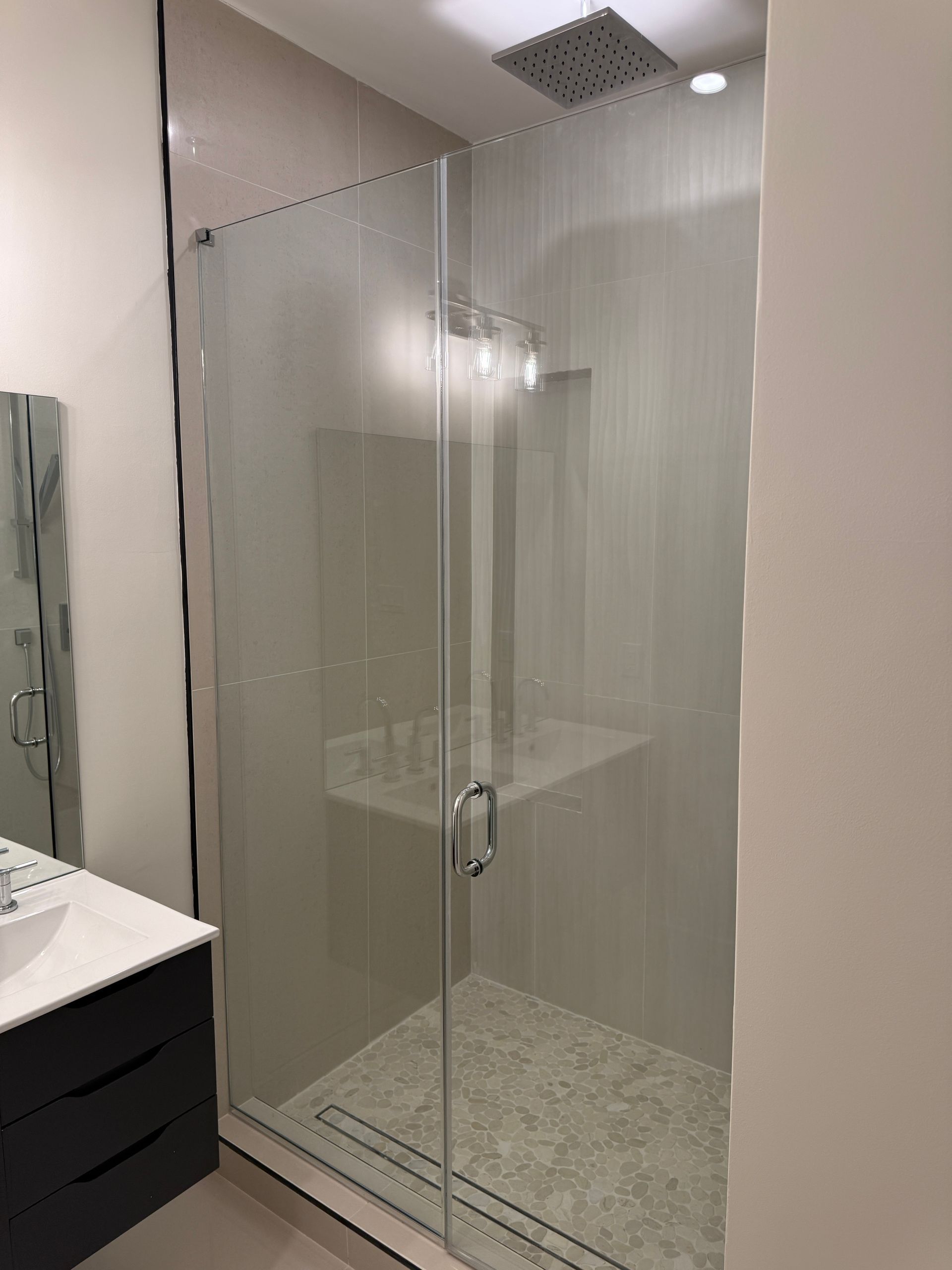 A frameless glass shower door with a silver handle installed in a modern bathroom with light tiles and a black vanity.