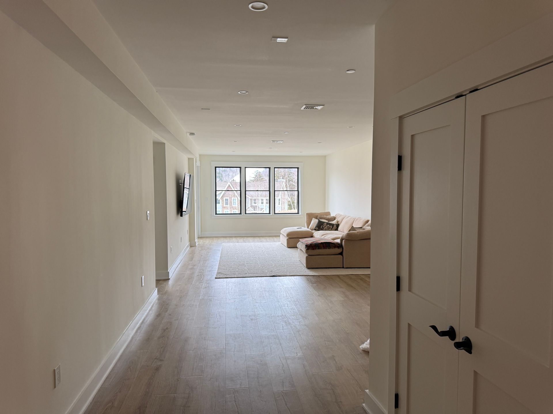 A long, open-concept living area with light wood floors, white walls, recessed lighting, and a beige sectional sofa.