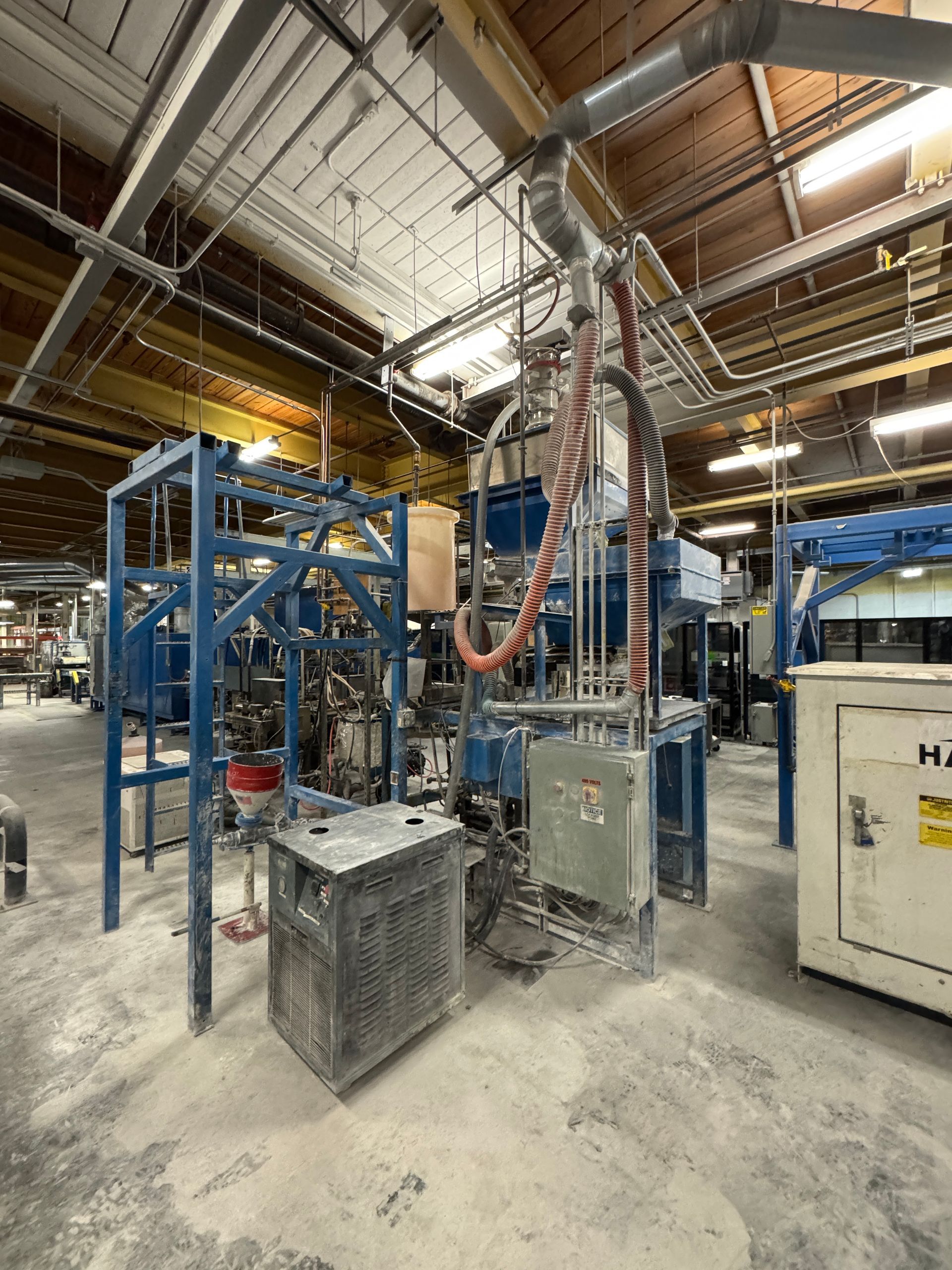Industrial machinery, various blue frames and piping, with a gray metal cage, inside a factory.