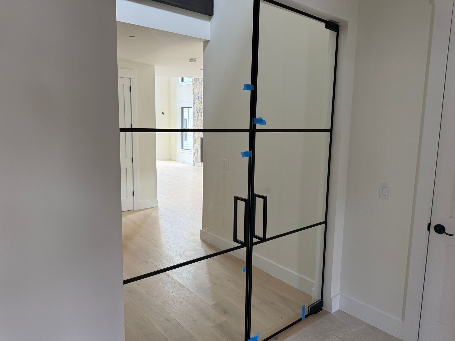 A pair of modern black-framed glass French doors with square handles installed in a brightly lit hallway.