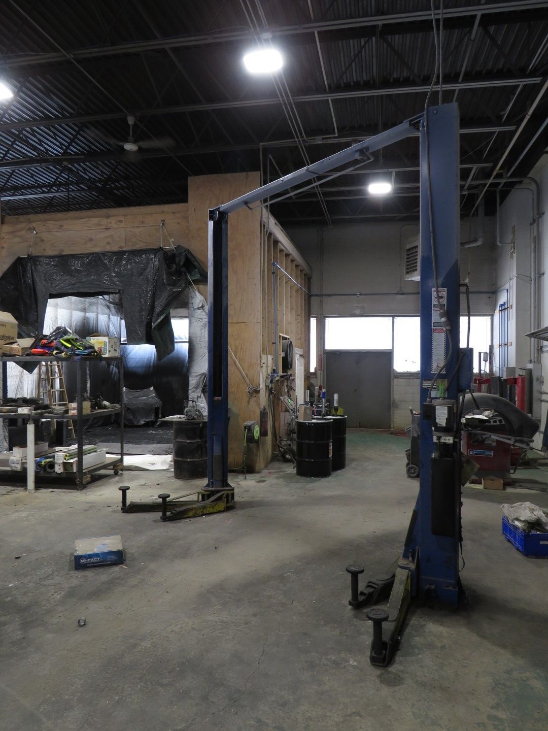 Two-post car lift in a workshop, concrete floor, overhead lighting, blue lift arms, tools and equipment visible.