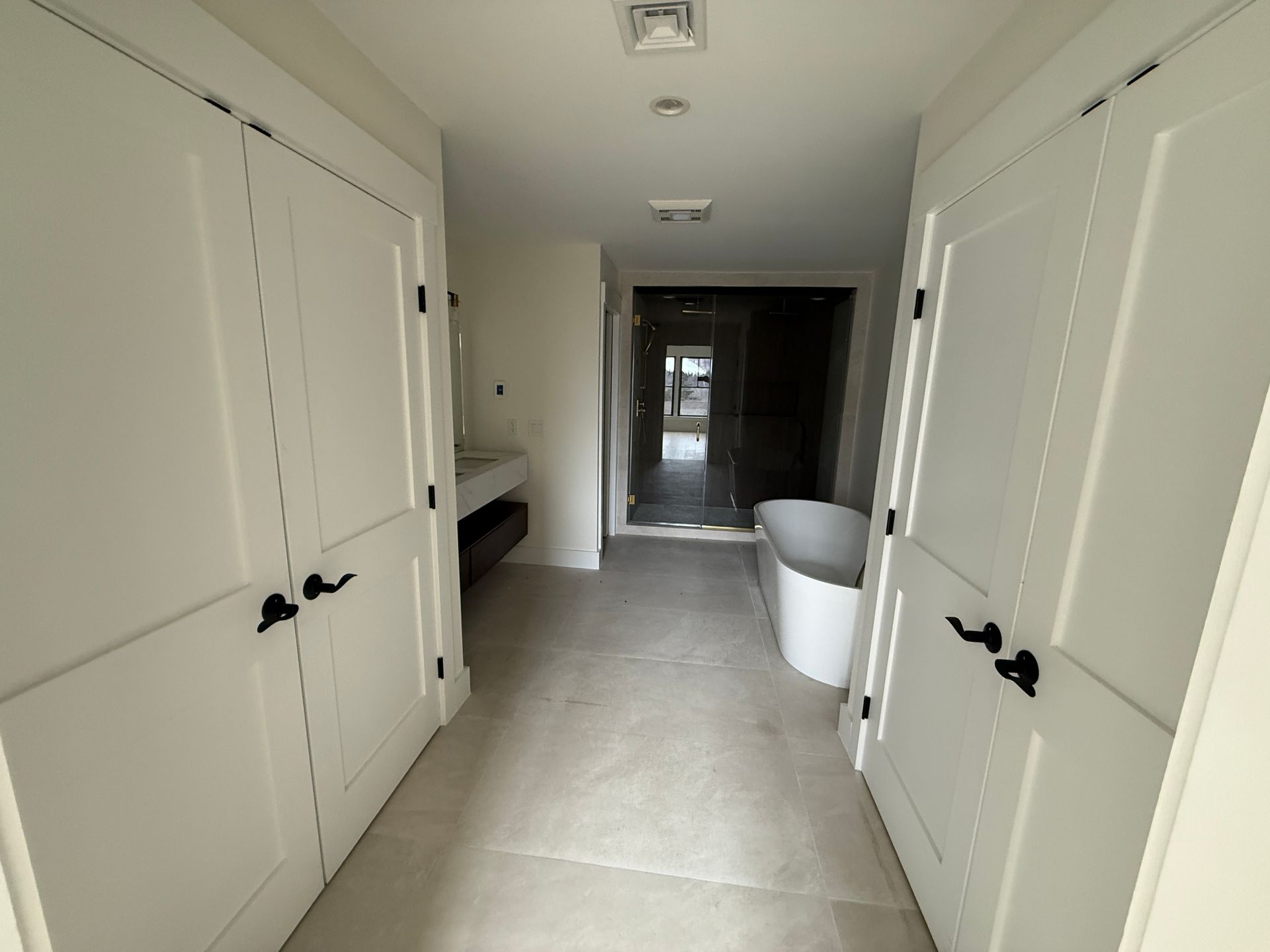 A hallway leads to a modern bathroom with a white freestanding tub, glass-enclosed shower, and white double doors.