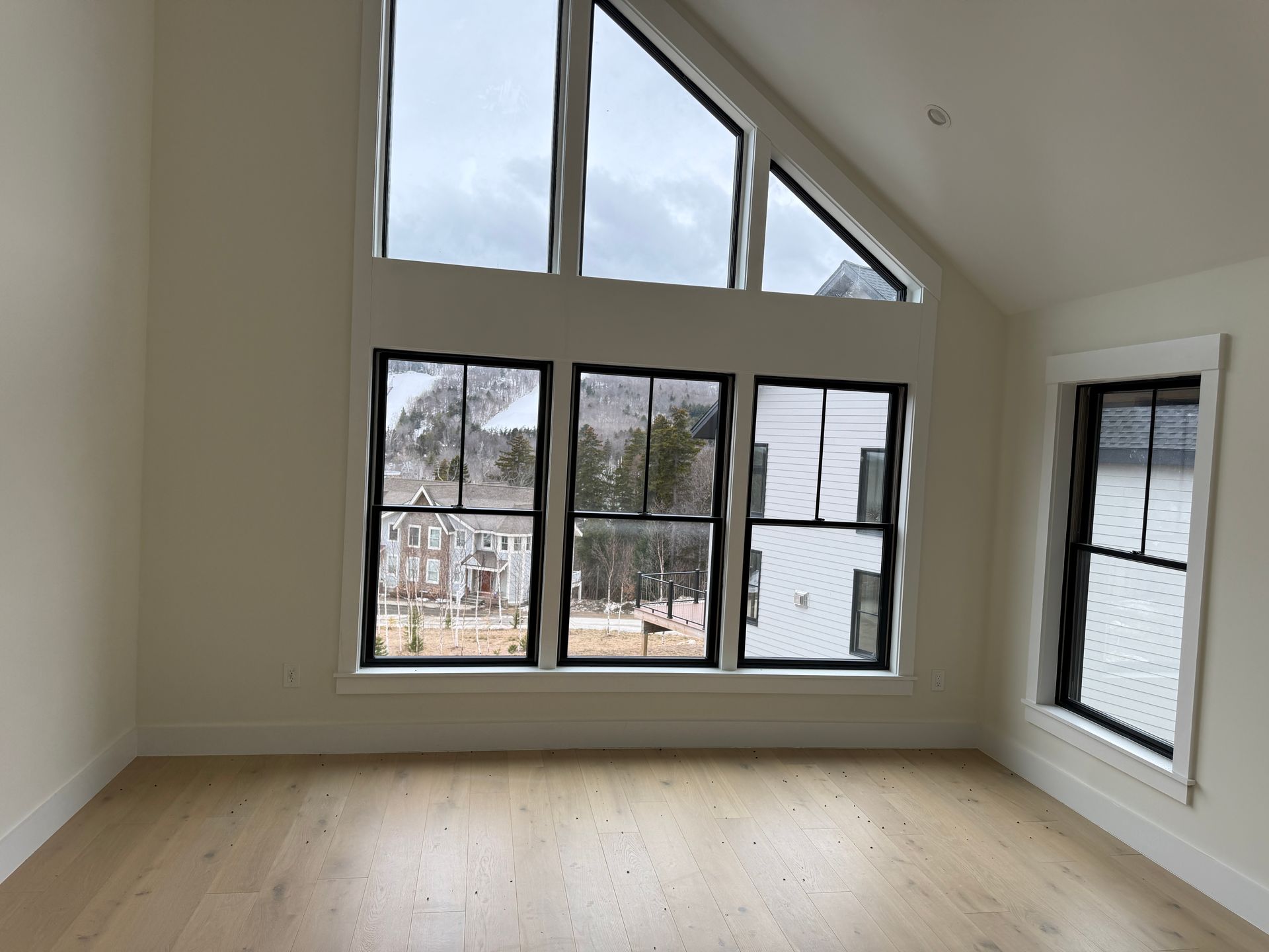 A spacious room with light hardwood floors, cream walls, and large windows with black frames looking out to a neighborhood.