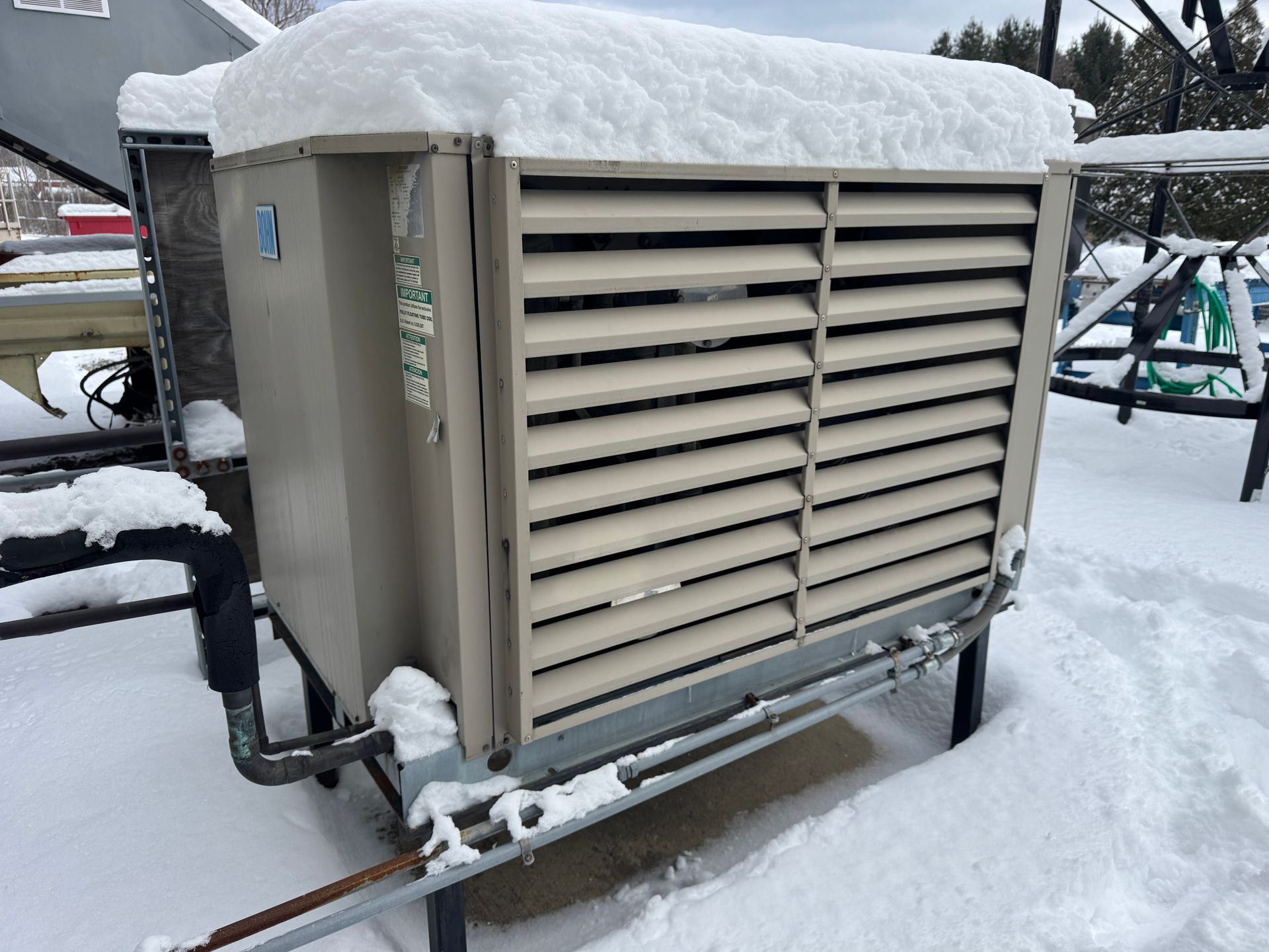 Snow-covered beige AC unit with horizontal slats, on a metal frame, in a snowy outdoor setting.
