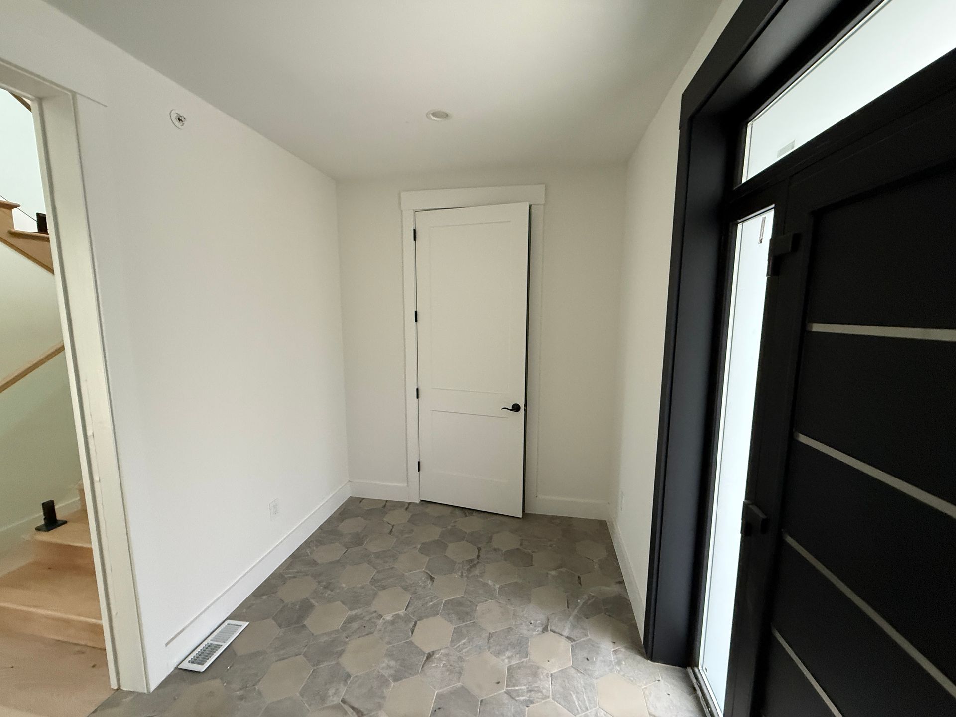 A brightly lit entryway with a dark door on the right, white walls, and light-colored hexagonal tile flooring.