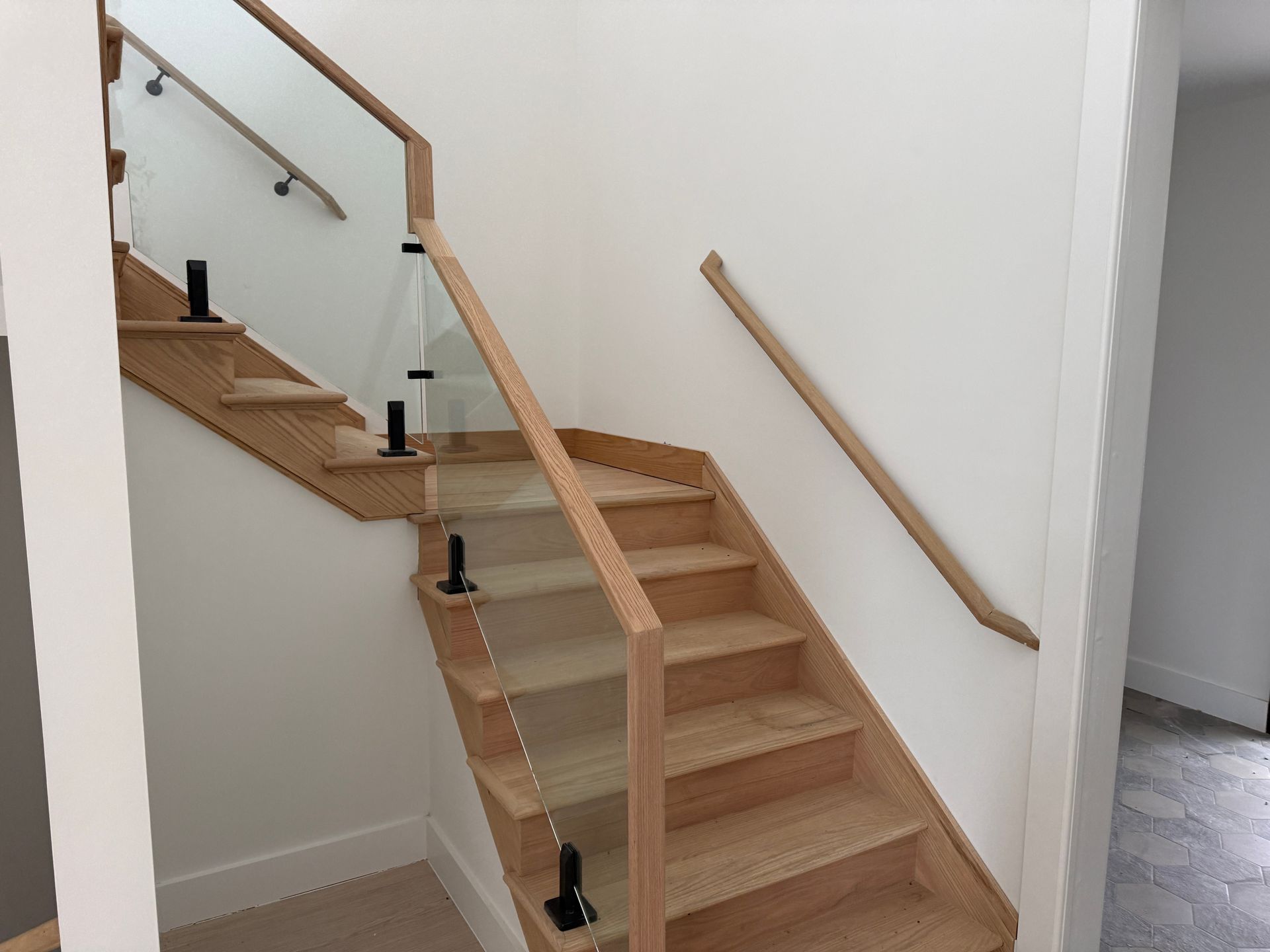 Light oak staircase with glass railings, black metal posts, and a matching wooden handrail on the adjacent wall.