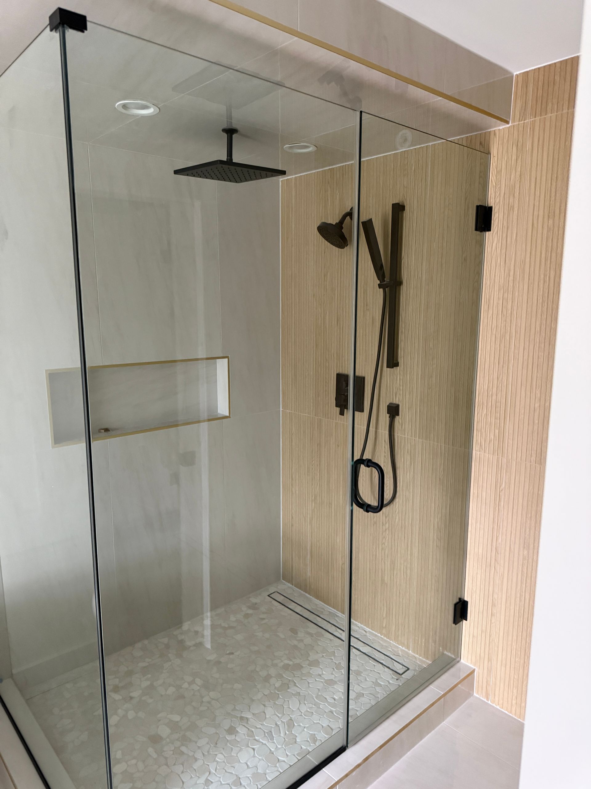 A modern walk-in shower with a rain showerhead, handheld sprayer, glass enclosure, and stone-tiled walls.