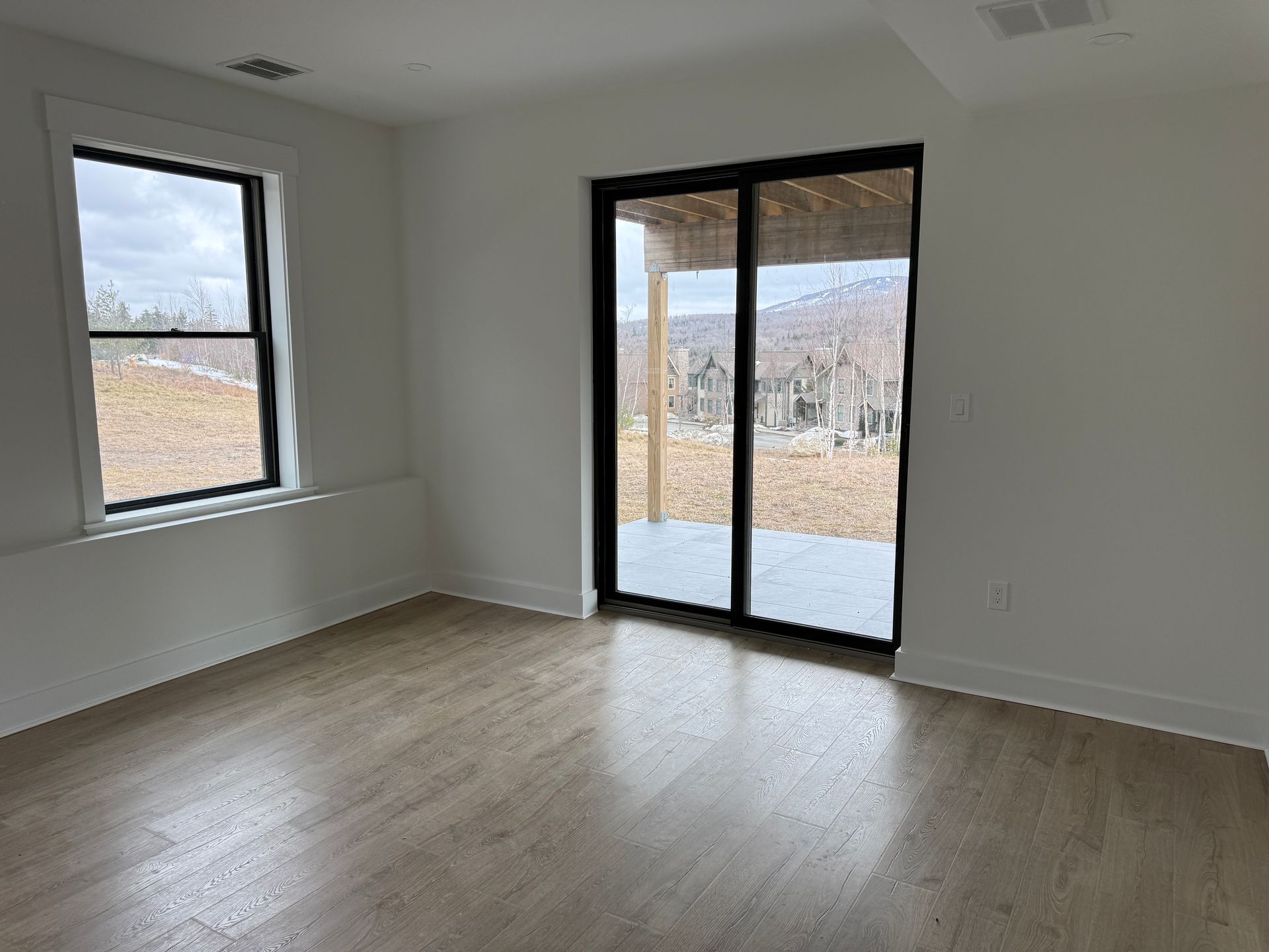 An empty, neutral-toned room with light wood floors, a single window, and a sliding glass door opening to a patio.