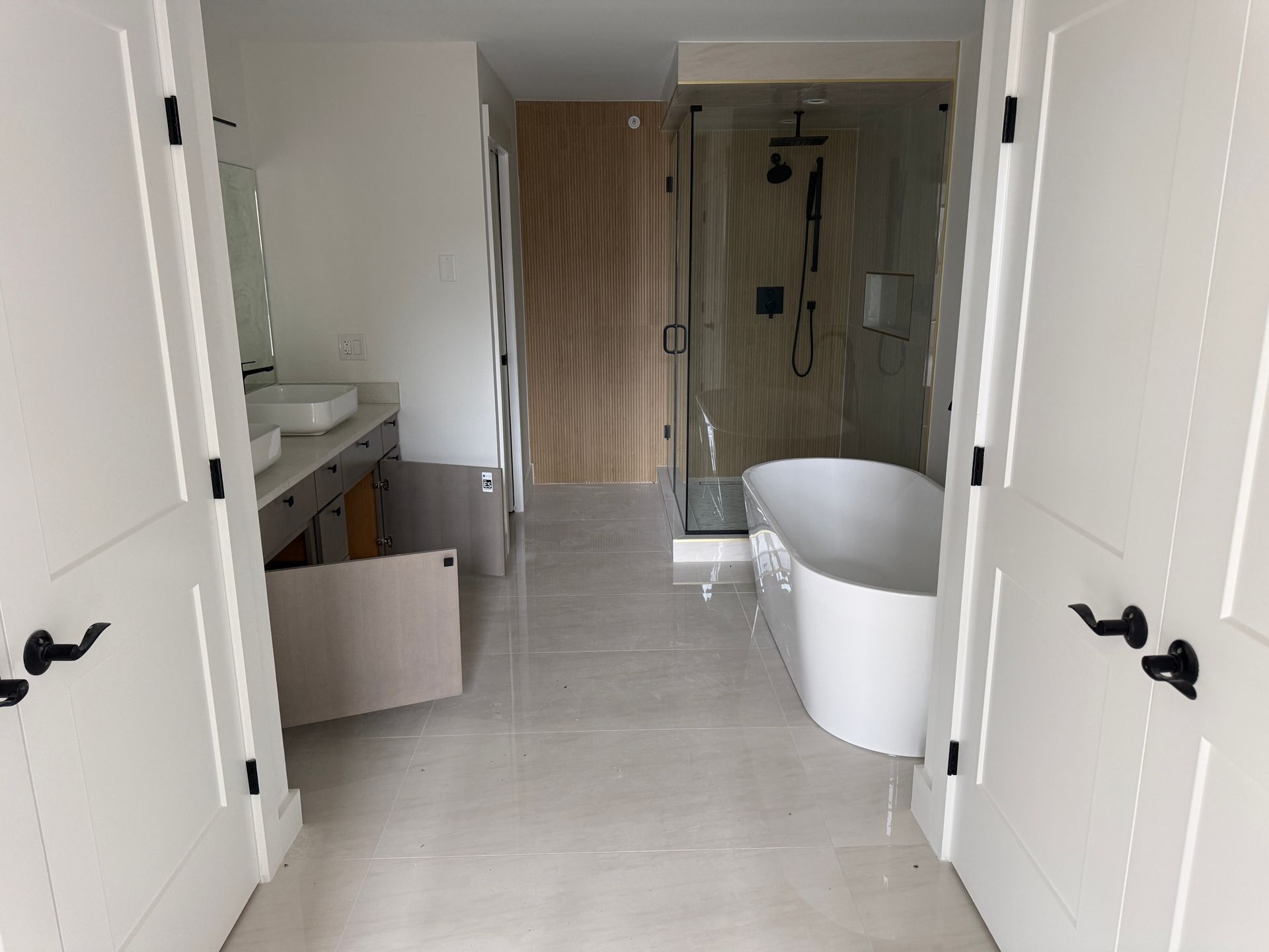 A modern bathroom featuring a white soaking tub, a glass-enclosed shower, dual vanity, and light wood door.