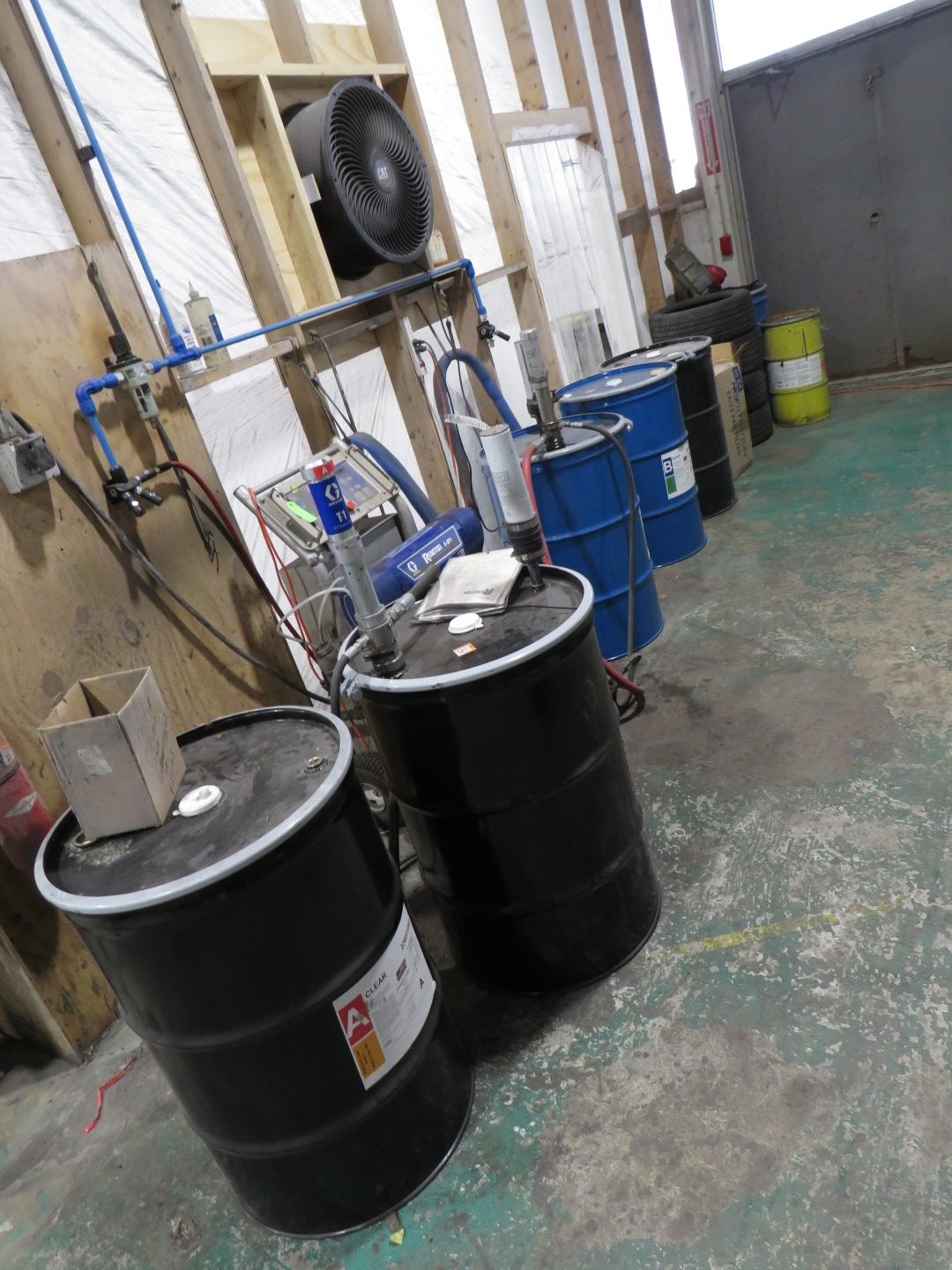 Rows of black and blue barrels in a workshop, some with equipment attached, against a wall with pipes and a fan.