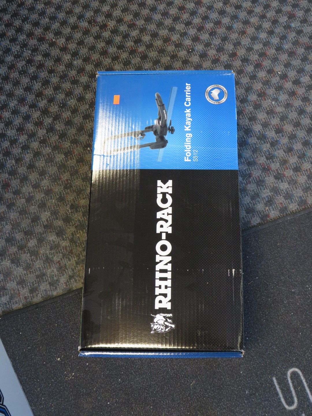 Rhino-Rack box, blue and black, for a folding kayak carrier, on a patterned surface.