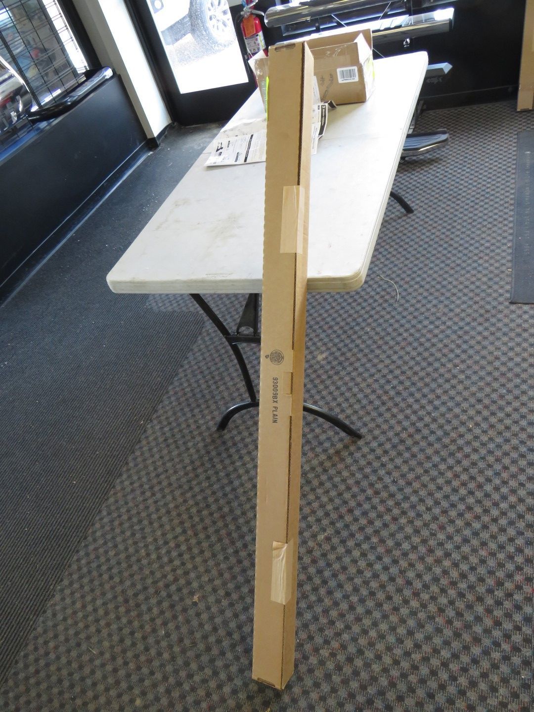 Tall cardboard box stands on a carpeted floor, near a folded white table with a box on it.