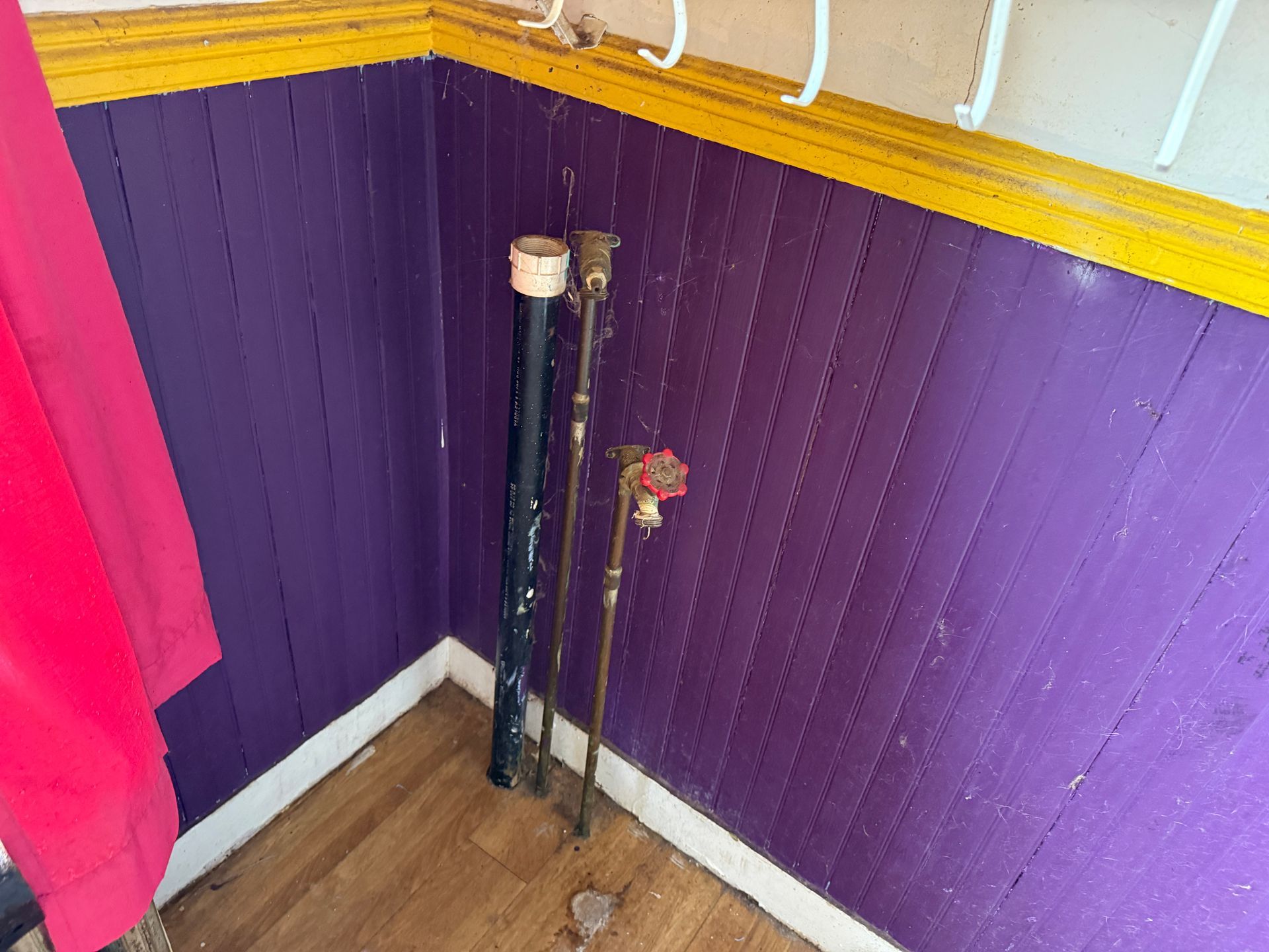 A corner with purple wooden paneled walls and yellow trim featuring two exposed pipes and a red valve on a wooden floor.