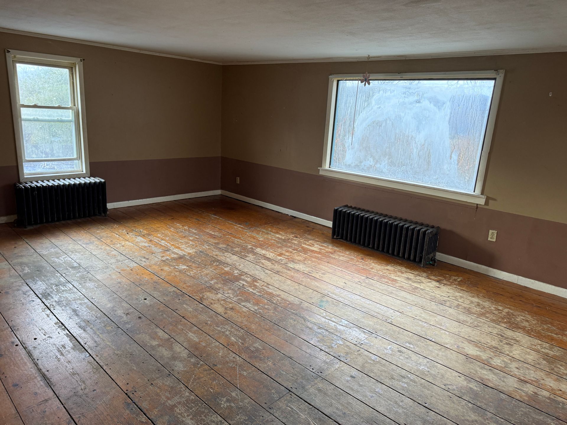 An empty room with light brown walls, dark wainscoting, two windows with black radiators, and wood plank floors.