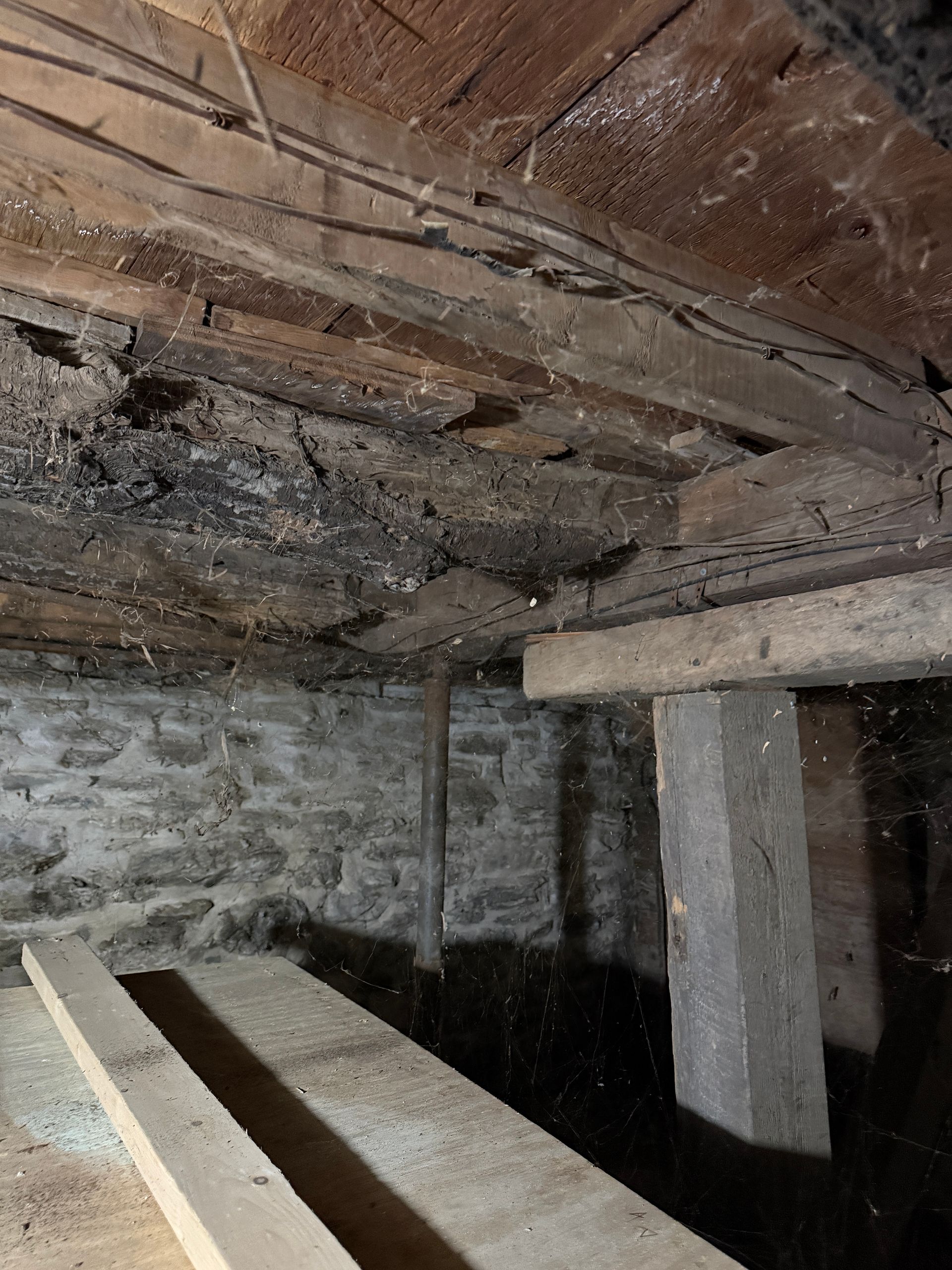 A low-angle view of a crawl space showing dark wooden floor joists, a stone wall, and a support post.