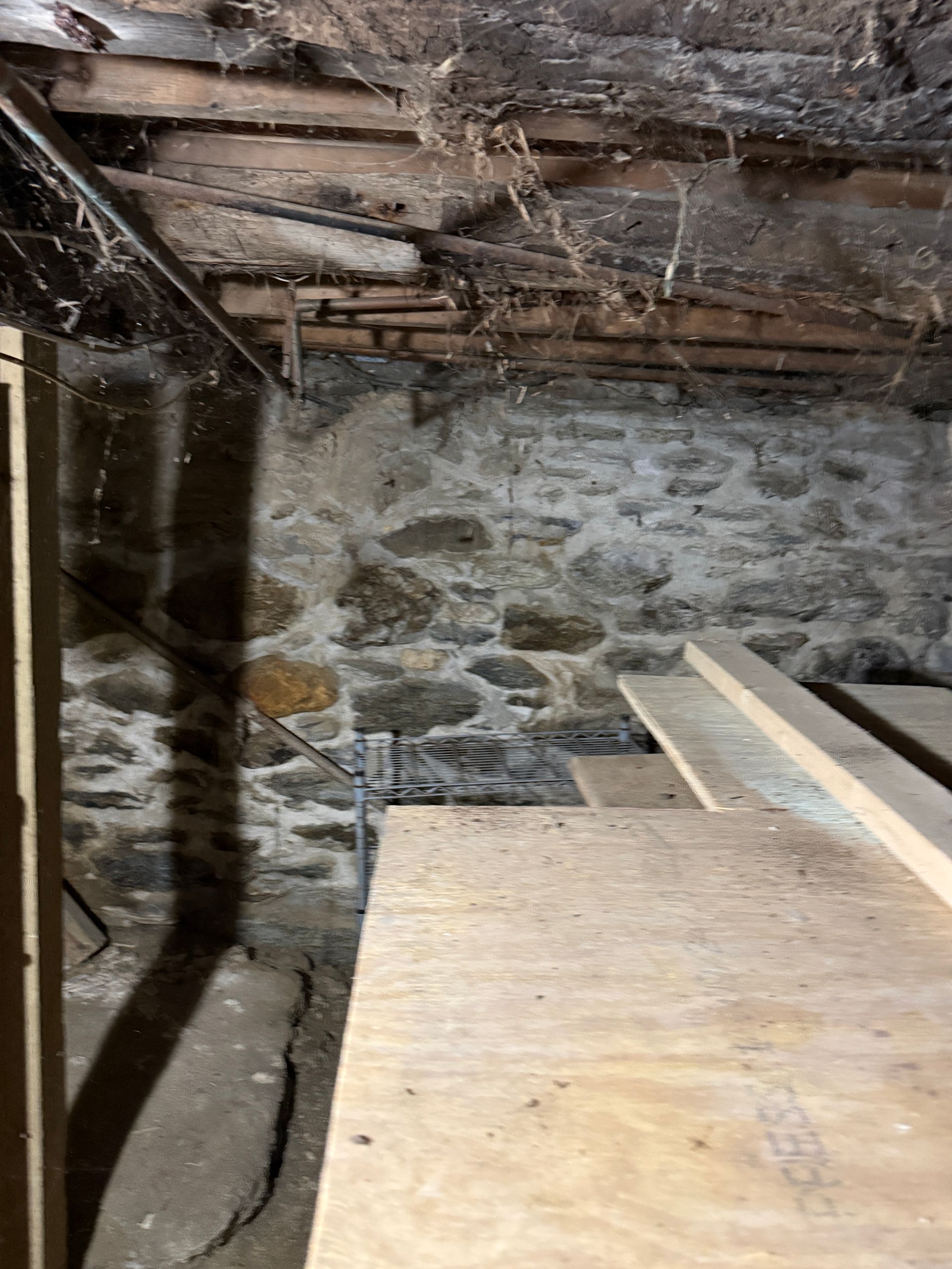 A low-angle view of a basement with a stone foundation wall, wooden ceiling joists, and plywood flooring.